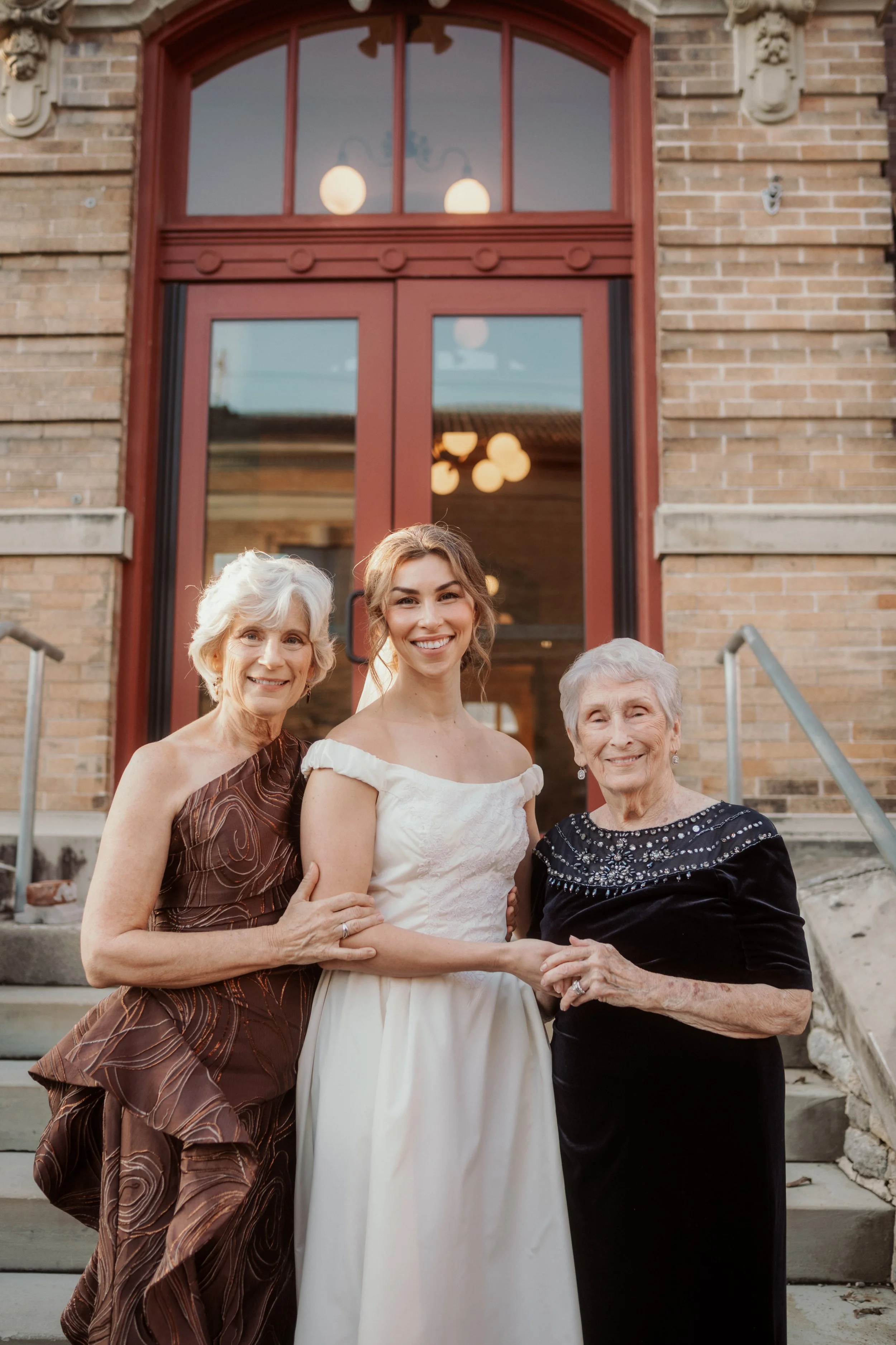 Four women standing on steps in front of a brick building with large window, smiling and holding hands, celebrating a wedding.