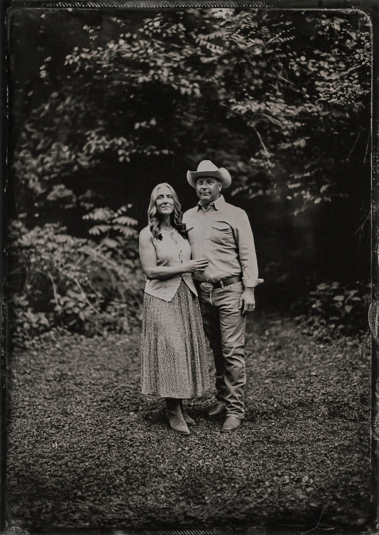 A black and white photograph of a man and woman standing outdoors, with trees and foliage in the background. The woman has long hair, is wearing a sleeveless top and a long patterned skirt, and is holding the man's arm. The man is wearing a cowboy ha