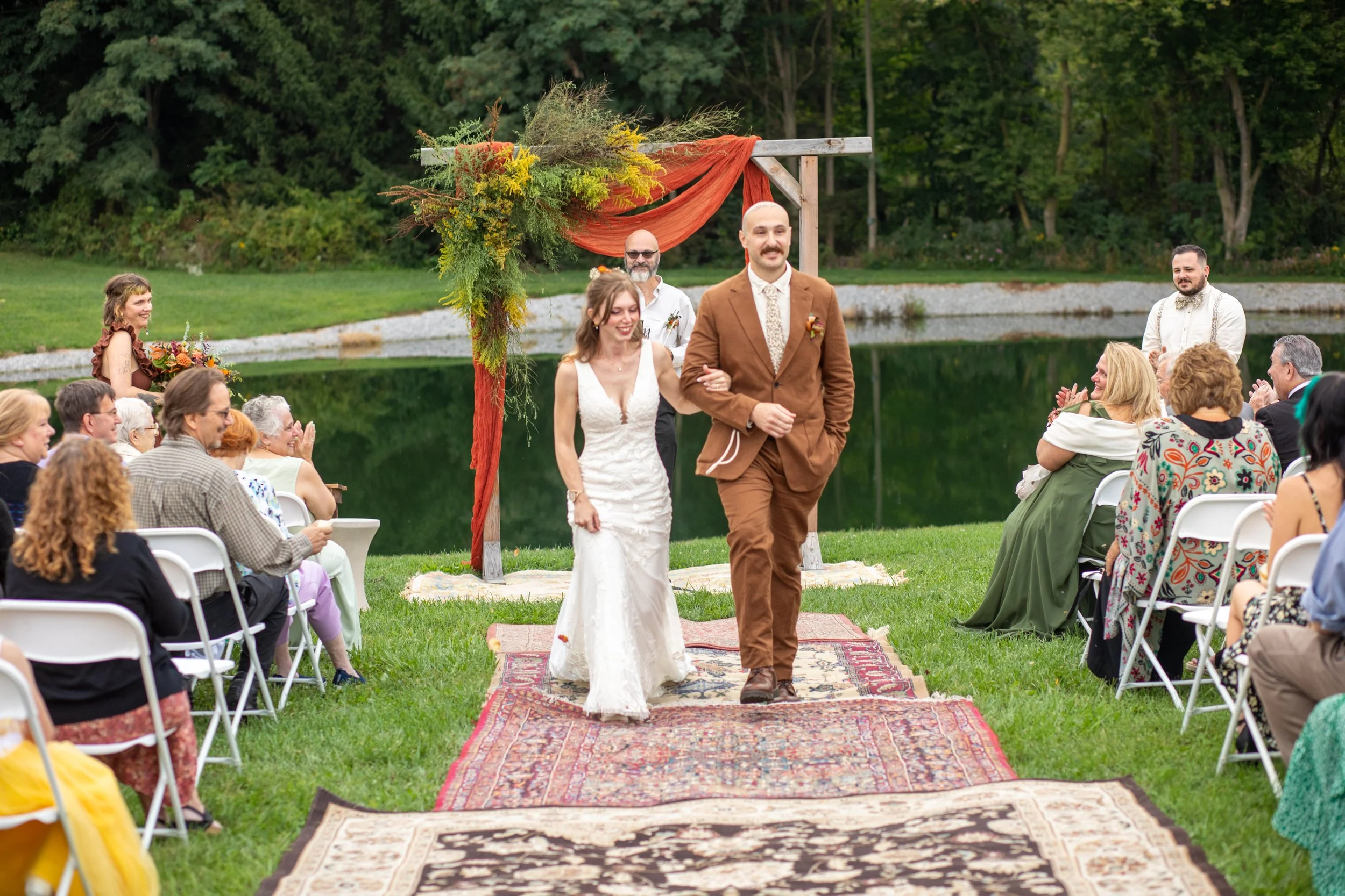 An outdoor wedding ceremony scene by a lake with a diverse group of guests seated in white chairs. A bride and groom walk down a red patterned aisle rug under a wooden arch decorated with greenery and an orange fabric. The scenic background includes 