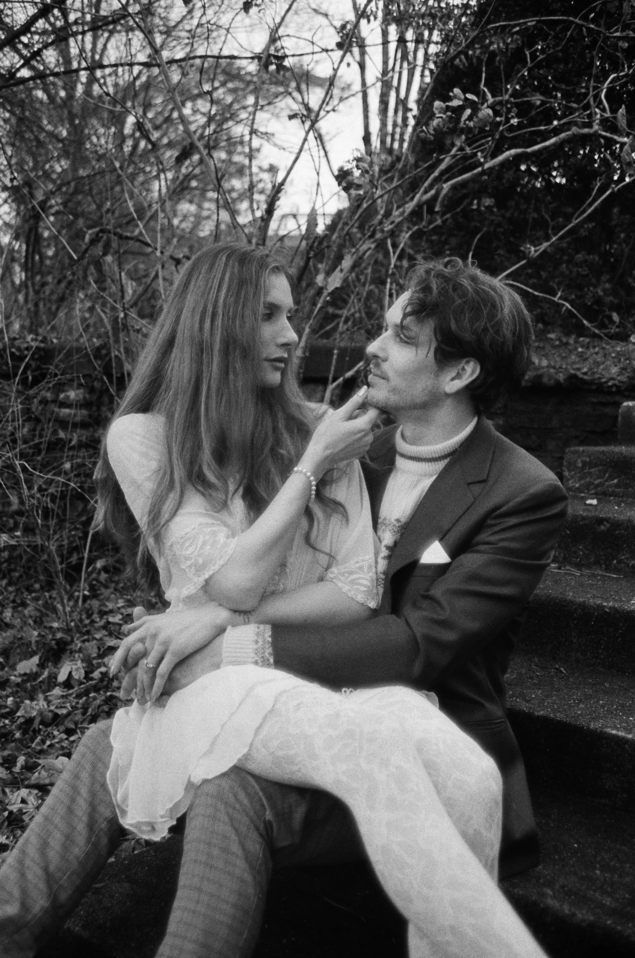 Black and white photo of a man and woman sitting outdoors among trees. The woman touches the man's chin while they look at each other.