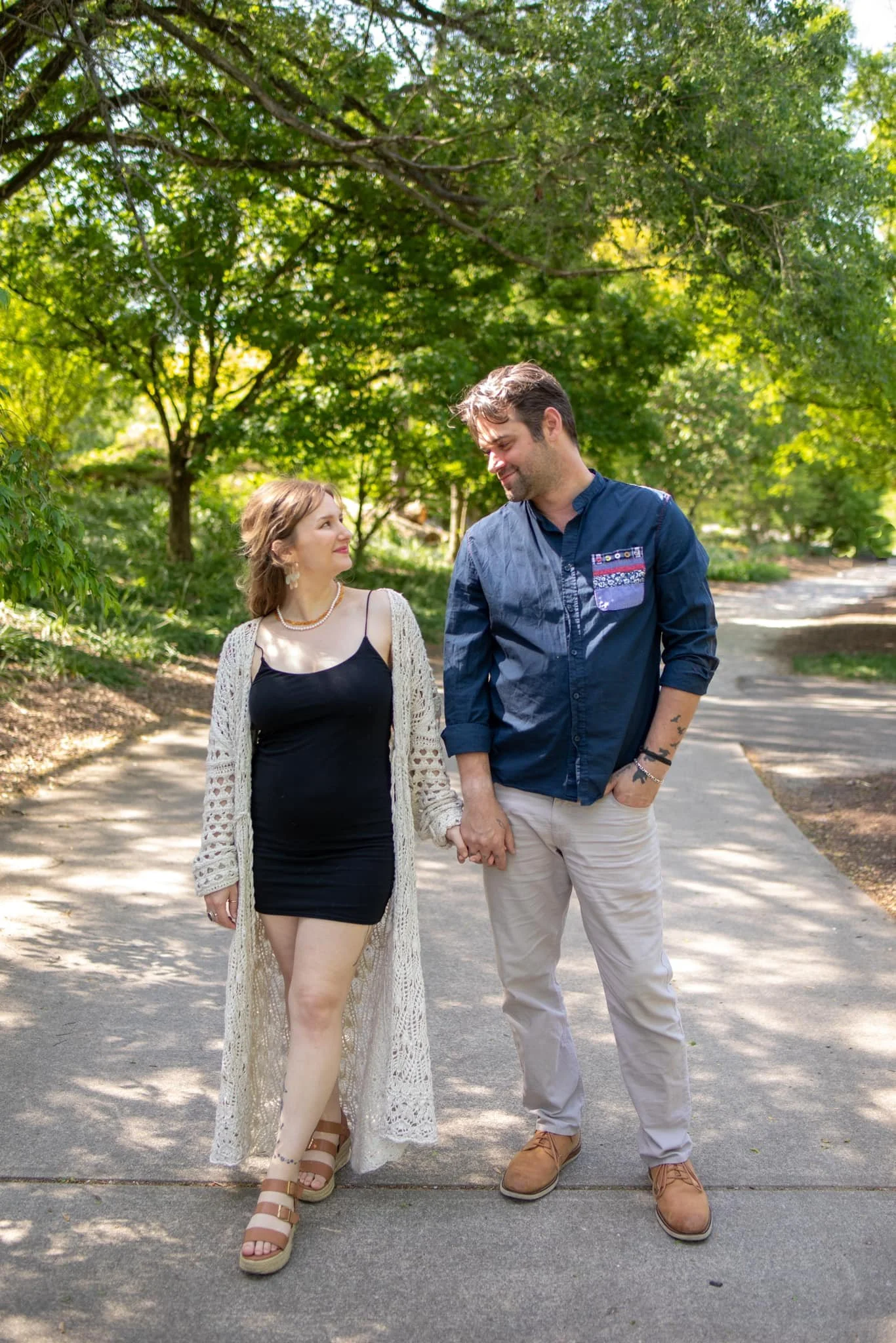 A couple holding hands walking on a sidewalk in a park with green trees overhead, smiling and looking into each other's eyes.