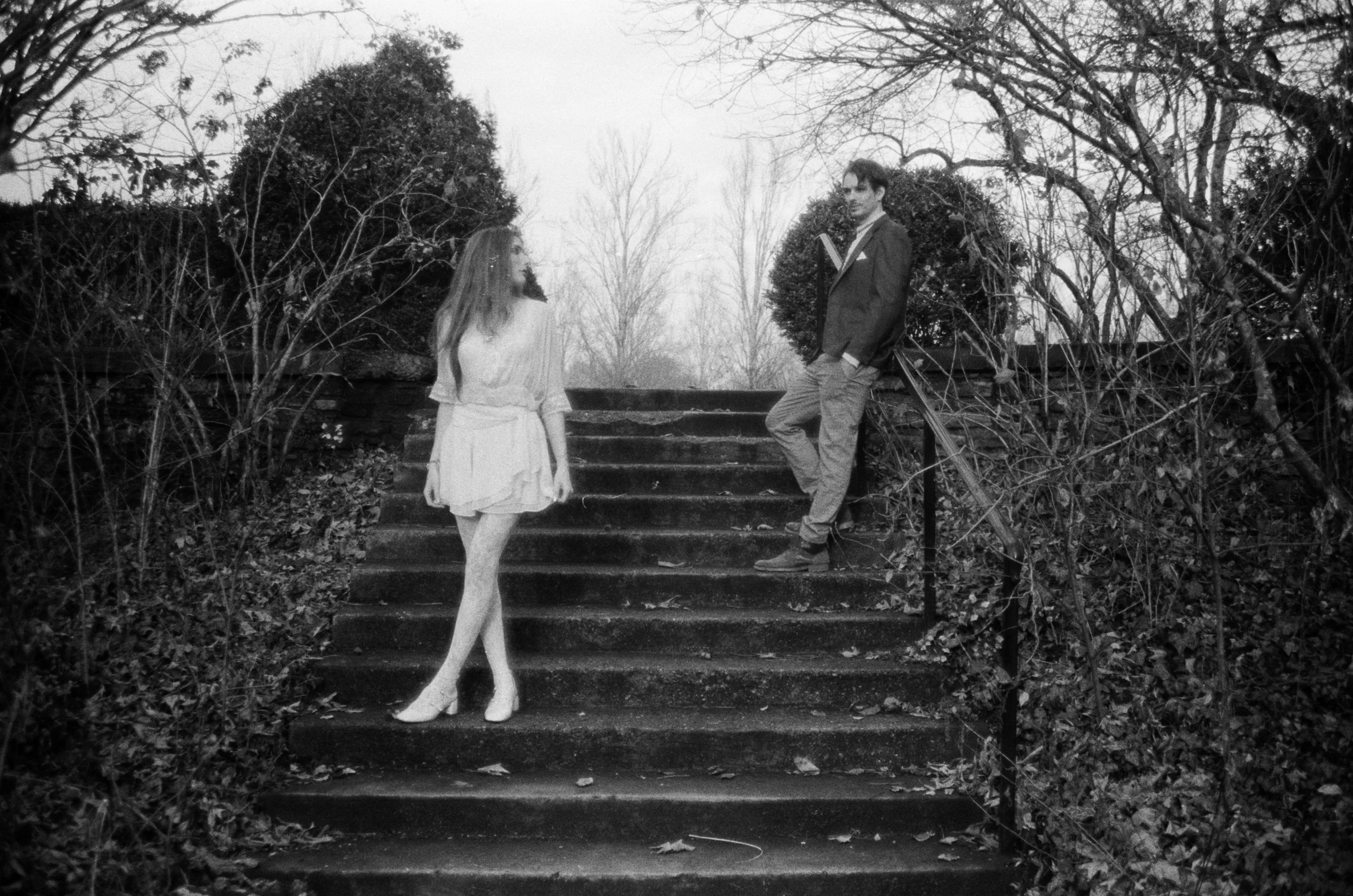 Black and white photo of a woman standing on stairs outdoors, facing towards a man who is leaning against a railing, surrounded by leafless trees and bushes.
