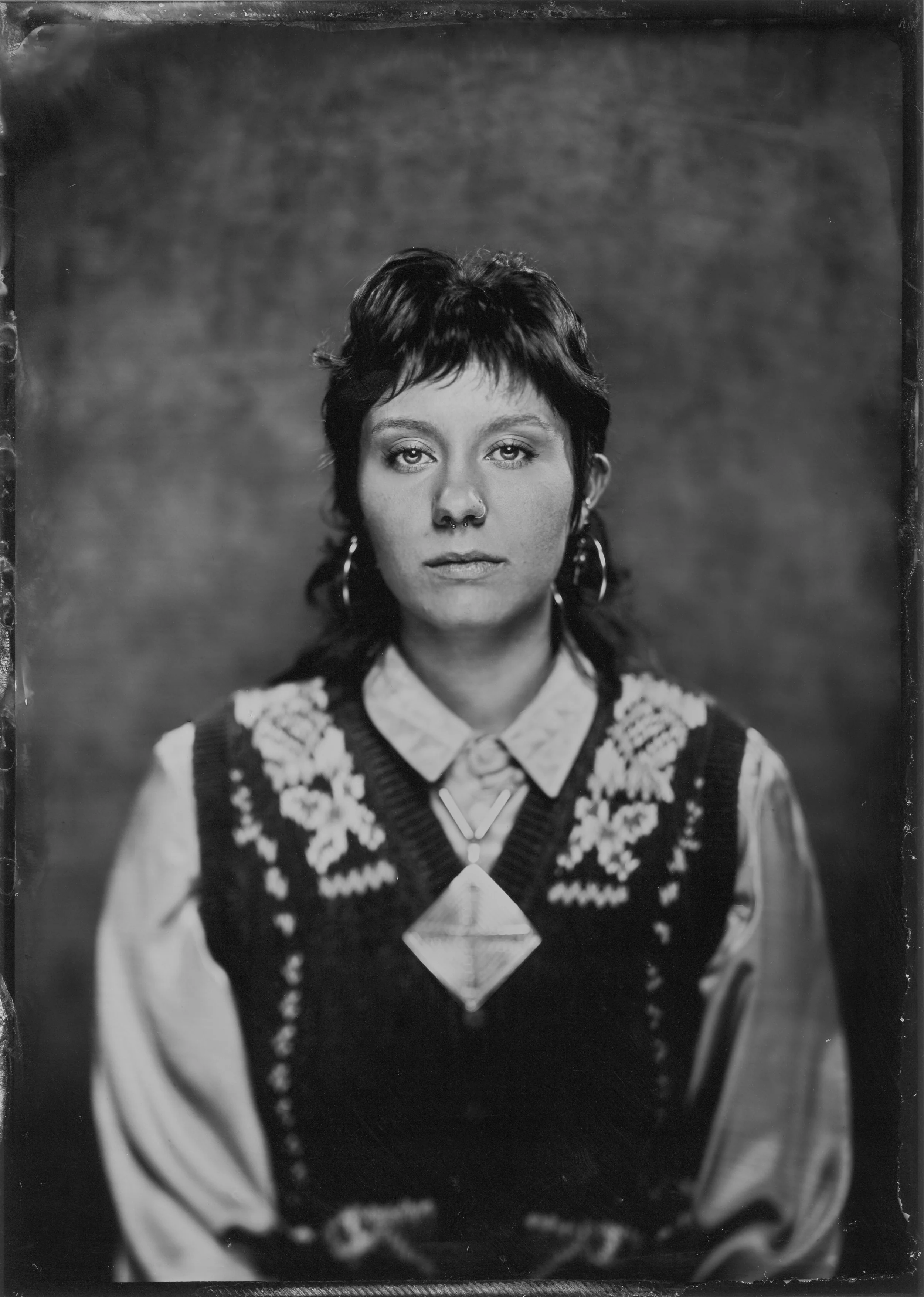 Black and white portrait of a woman with short hair, wearing hoop earrings, a collared shirt, and a patterned sweater vest, looking directly at the camera.