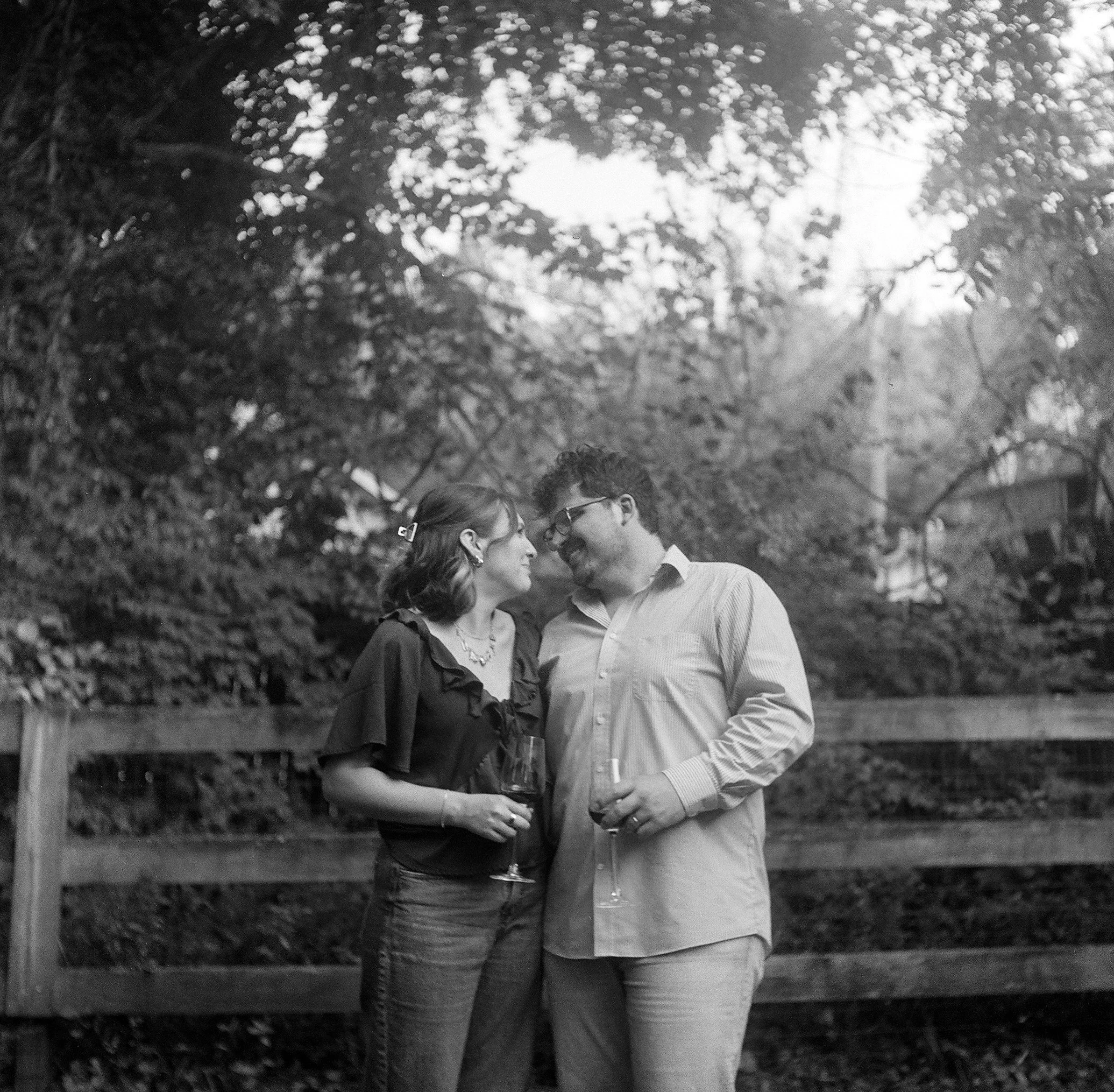 A black and white photograph of a couple standing close together outdoors, holding wine glasses, with trees and a fence in the background.