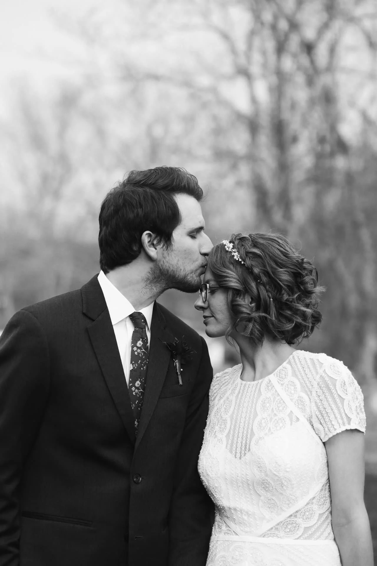 A black and white photograph of a bride and groom on their wedding day. The groom is kissing the bride on the forehead while both have their eyes closed, standing close together outdoors with trees in the background. The groom is dressed in a suit wi