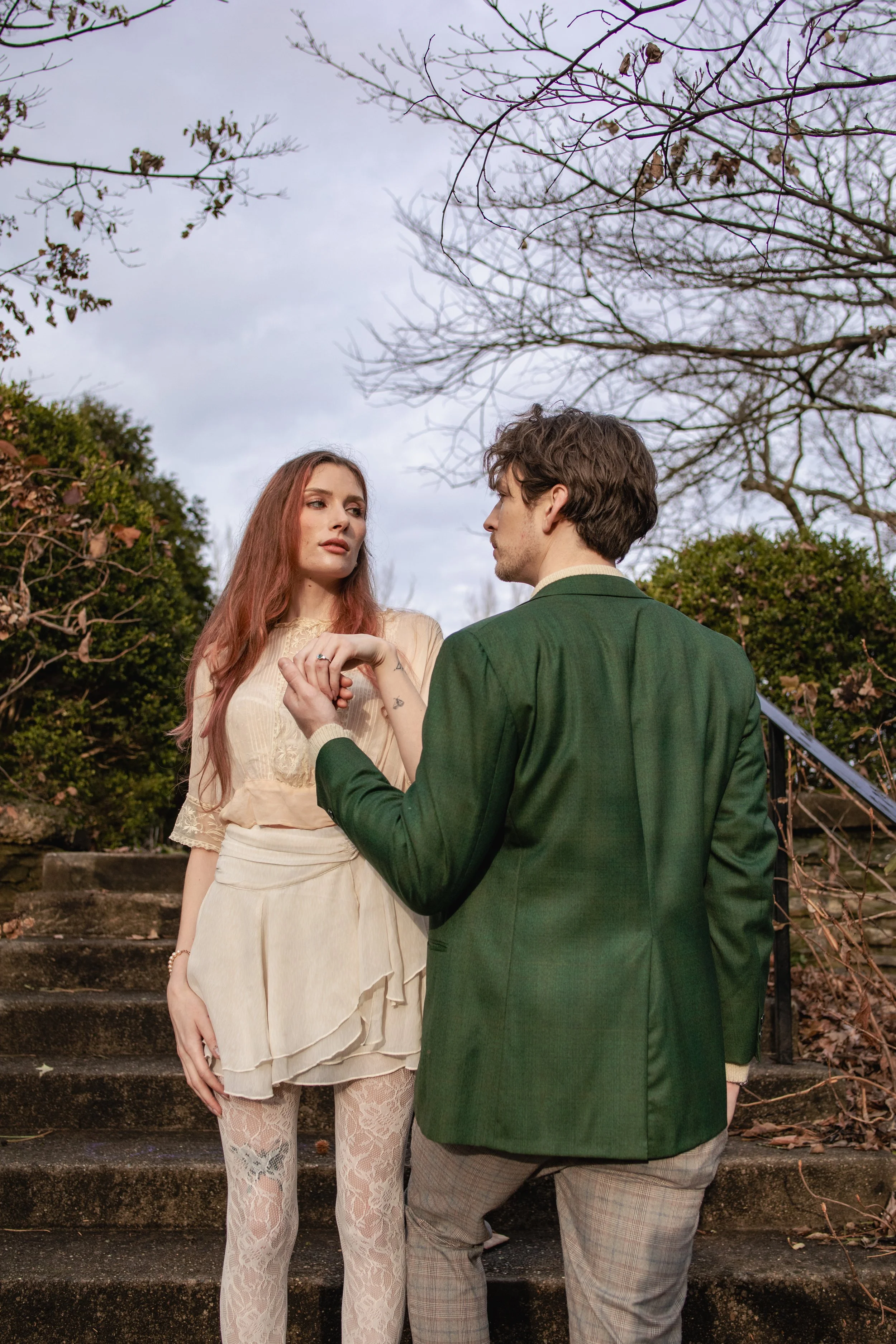 A young woman with long red hair and patterned lace pants is holding hands with a young man in a green blazer and plaid pants while standing on outdoor stone steps surrounded by leafless trees and bushes.