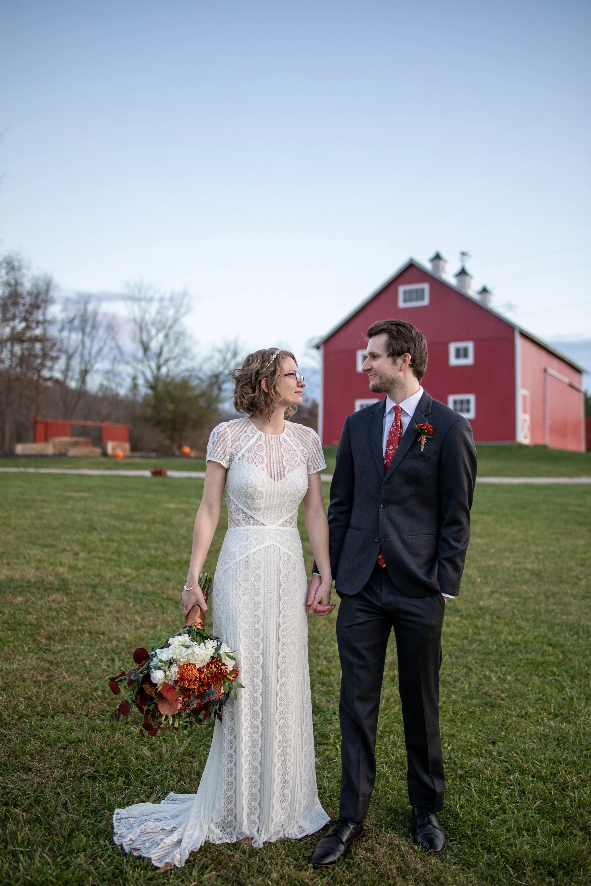 A bride and groom holding hands and smiling at each other outdoors, with a red barn in the background and cloudy sky overhead.