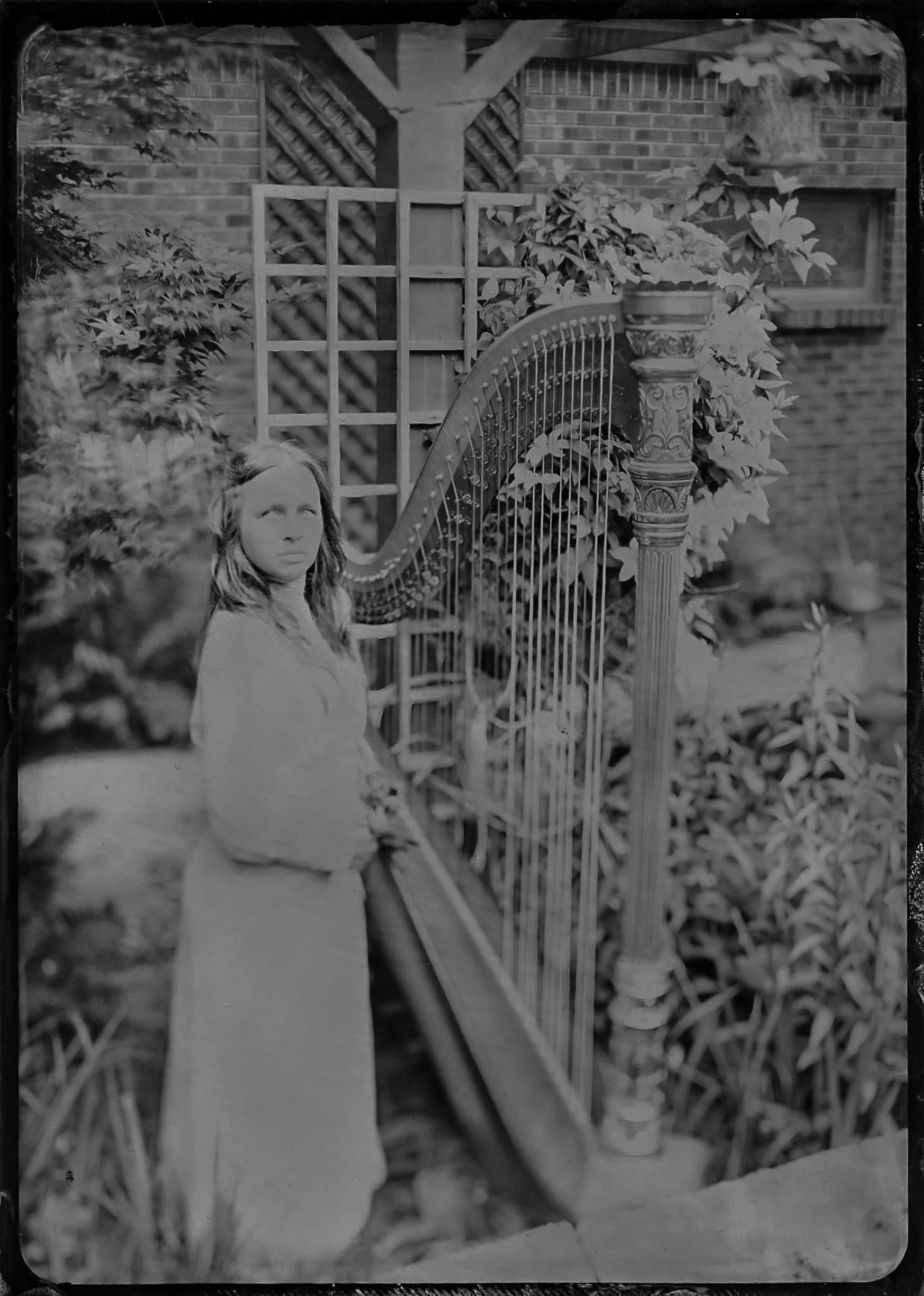 A woman with long hair wearing a coat sitting on a garden bench. Behind her, there is a trellis, a decorative column with a potted plant, and a brick house with a window. The garden has various plants and foliage.