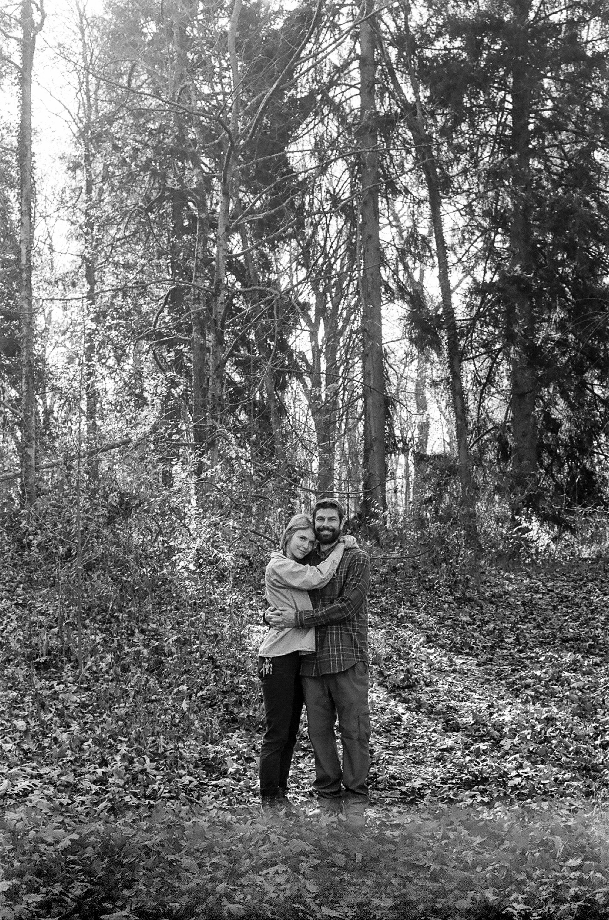 A black and white photo of a couple hugging in a woodland area with trees and fall foliage.