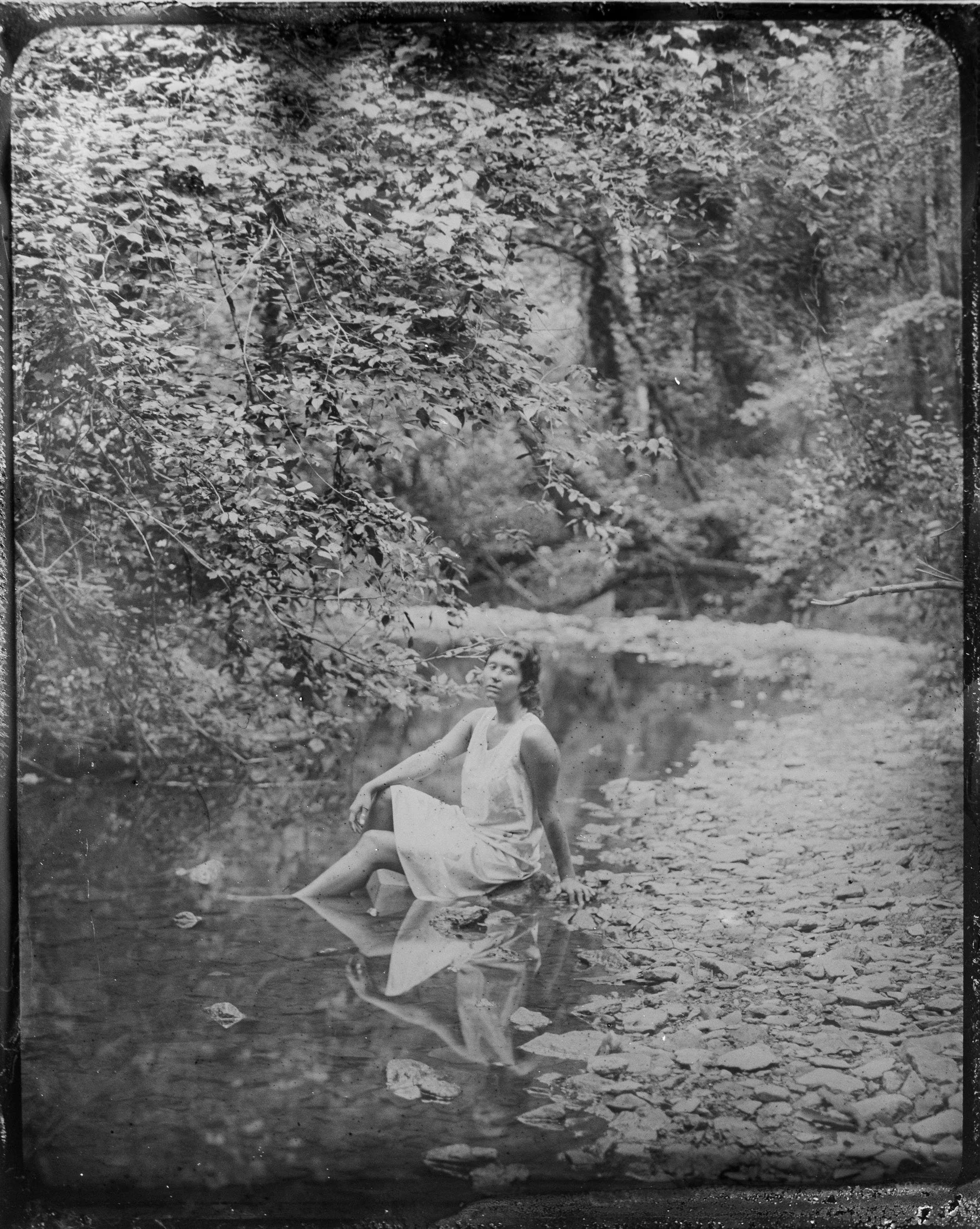 A black and white photograph of a woman sitting in a shallow stream in a forested area. She is wearing a light-colored dress and appears to be looking upward. The surrounding environment includes dense foliage and trees.