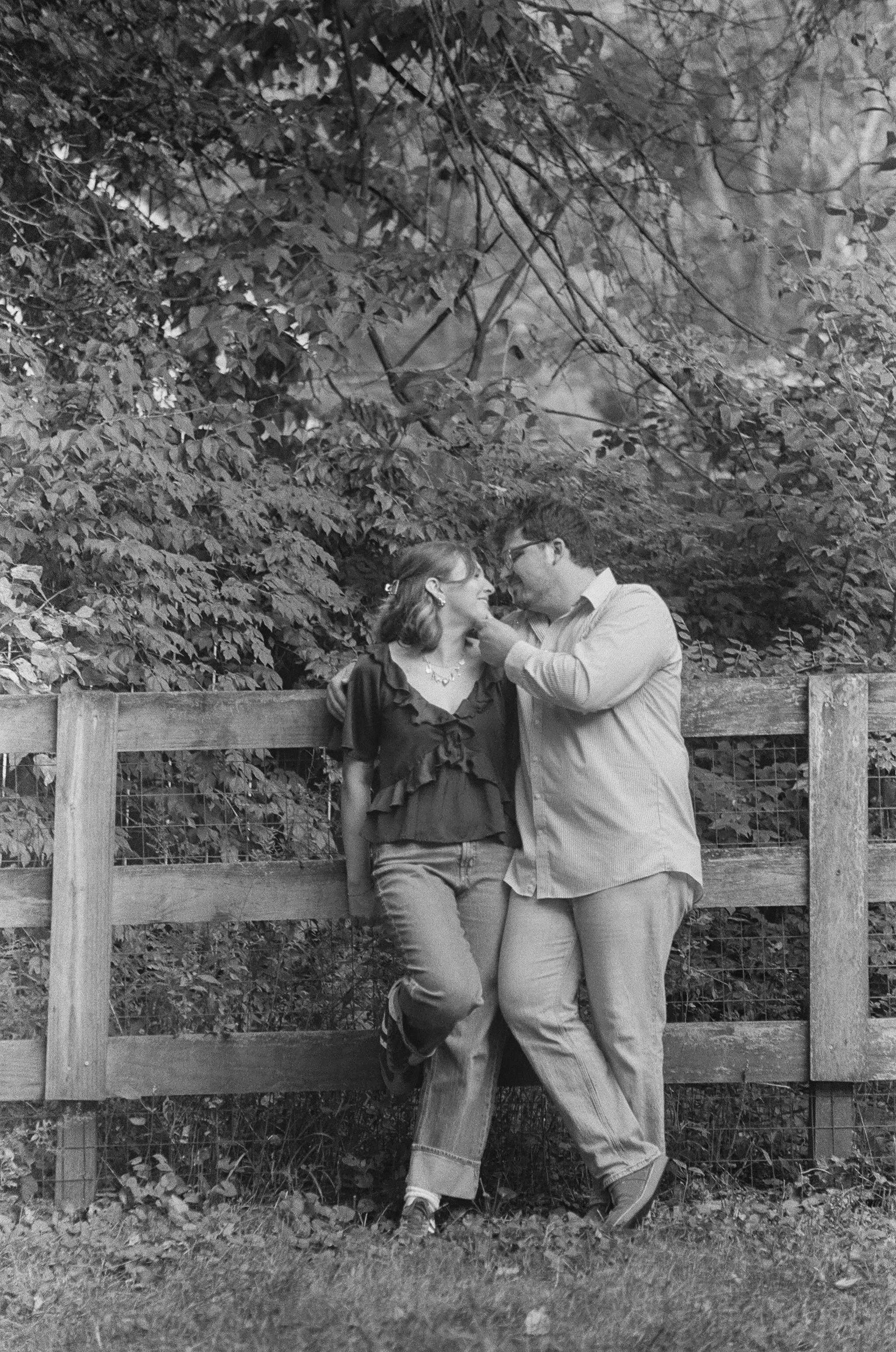 A black and white photo of a couple walking along a wooden fence with dense foliage in the background. The woman is wearing a blouse and jeans, and the man is dressed in a shirt and pants. They are touching foreheads and smiling at each other.
