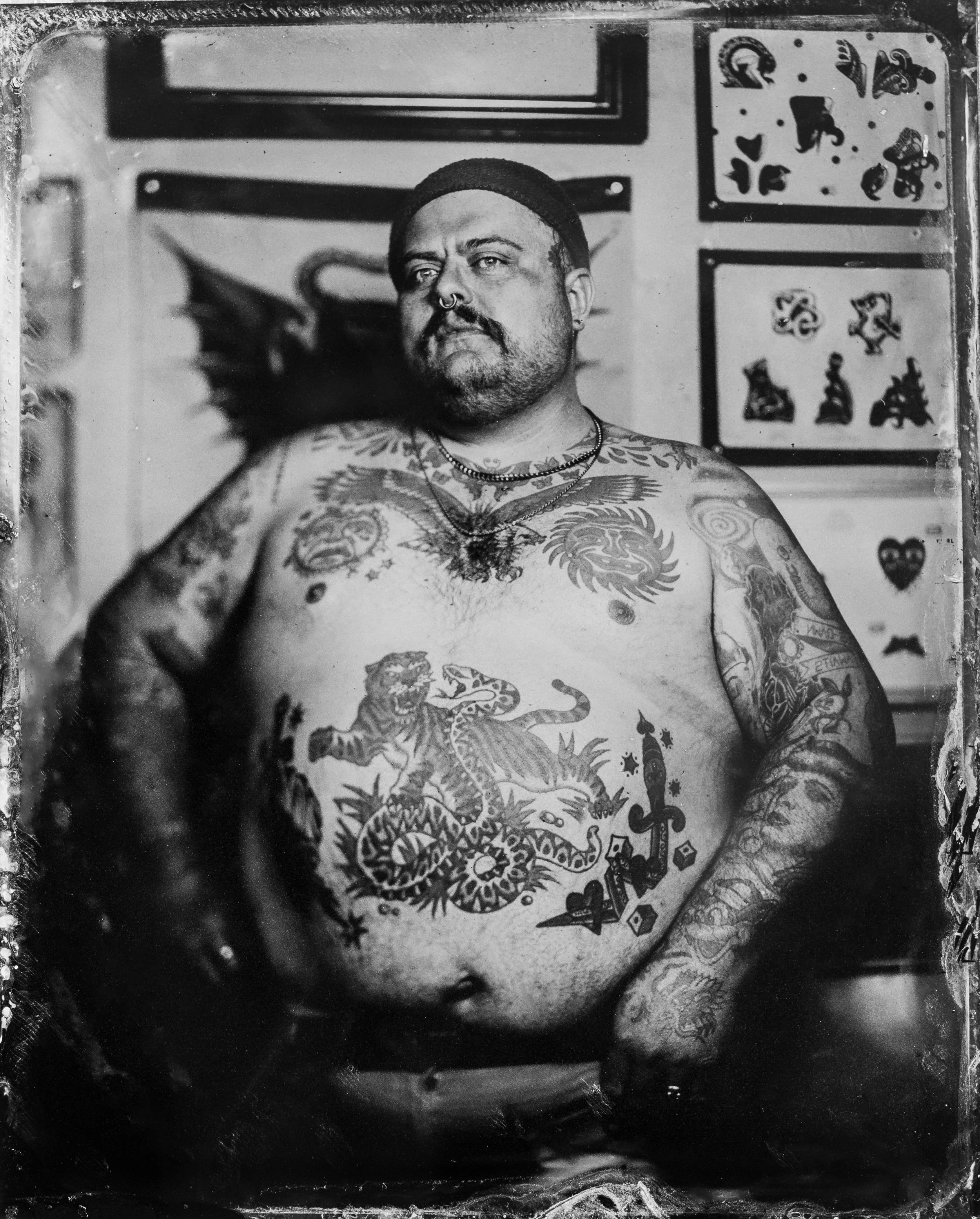 Black and white photograph of a tattooed man with tattoos of a lion and a snake on his chest, various tattoos on his arms, a pierced nose, and wearing chains, standing indoors with artwork on the walls behind him.