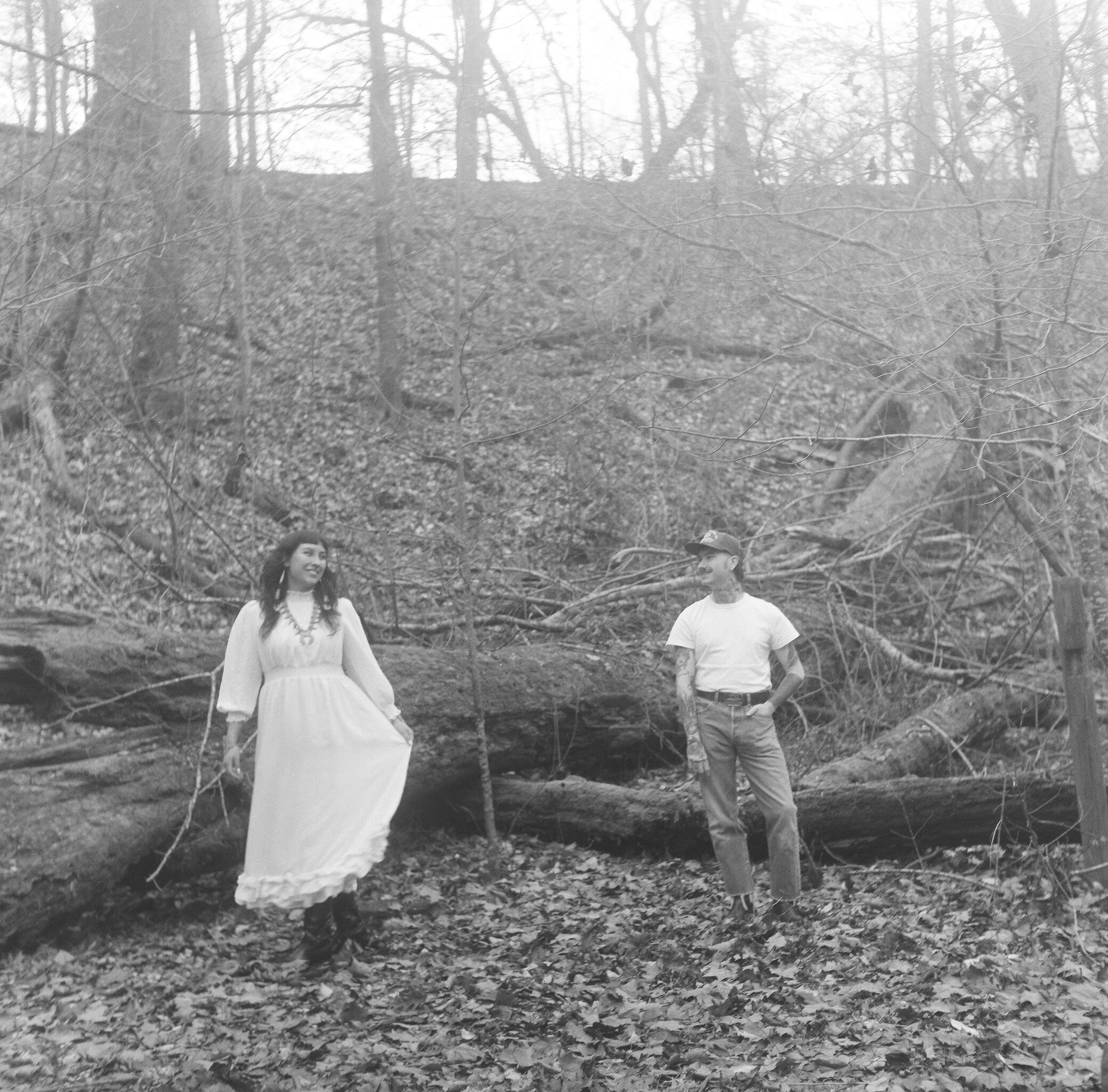 Black and white photograph of two young people, a woman and a man, standing in a wooded area with fallen leaves.