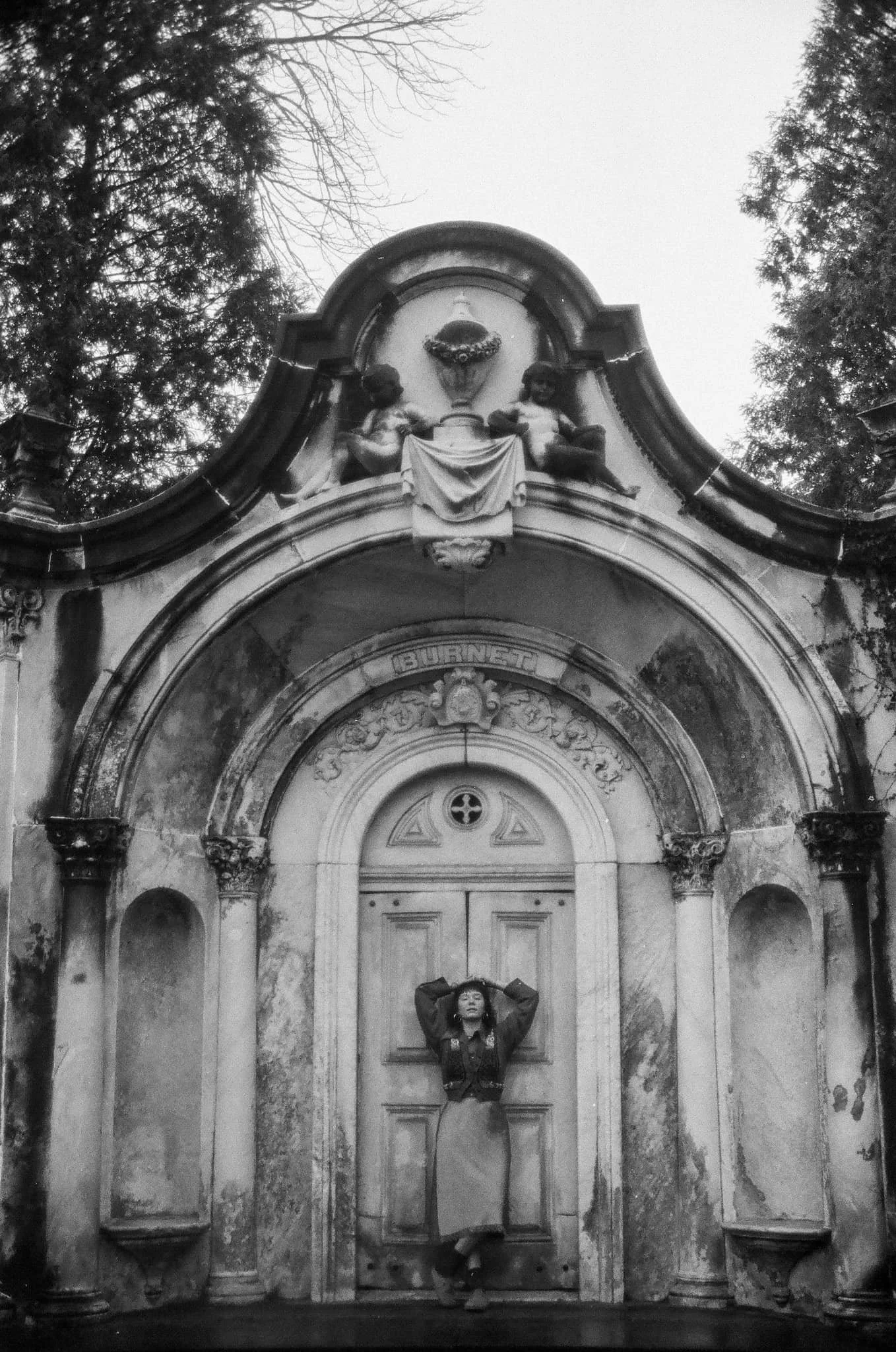 A woman stands in front of a vintage building entrance, posing with her hands behind her head. The building has intricate architectural details, including a sculpted relief of two children holding a shield or wreath, with trees visible in the backgro