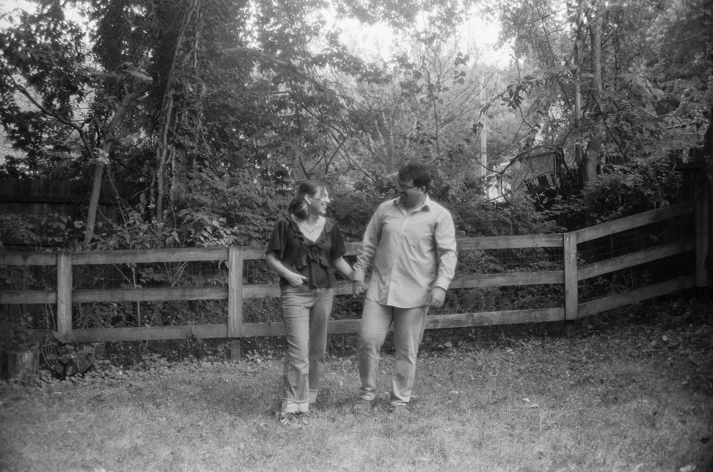 Two people, a woman and a man, walking hand in hand in a backyard with trees and a wooden fence.