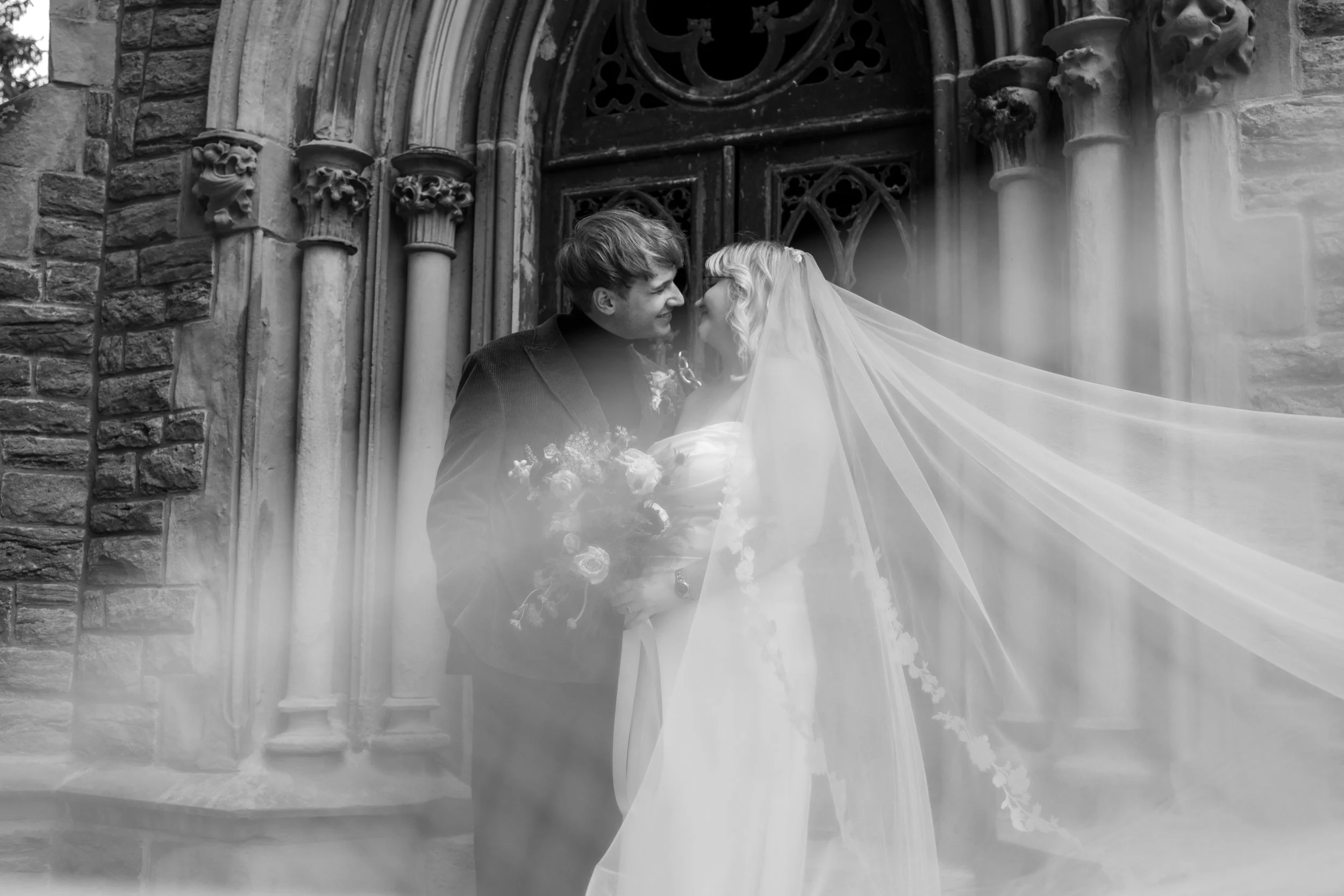 A black and white photo of a bride and groom smiling and leaning close to each other outside a church or historic building, with gothic architectural details and stone columns.