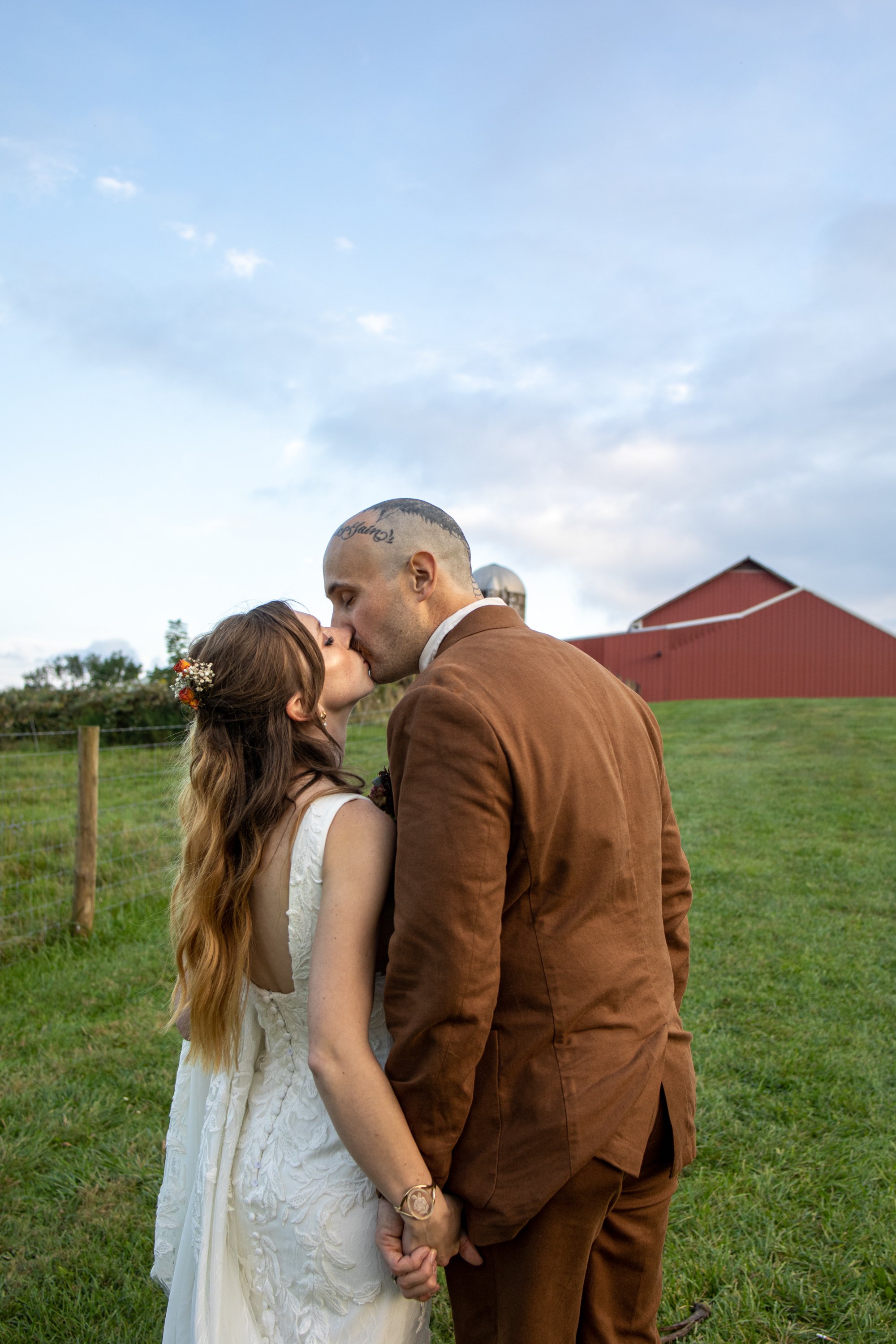 A couple kissing outdoors during what appears to be a wedding or special occasion, with a red barn and cloudy sky in the background.