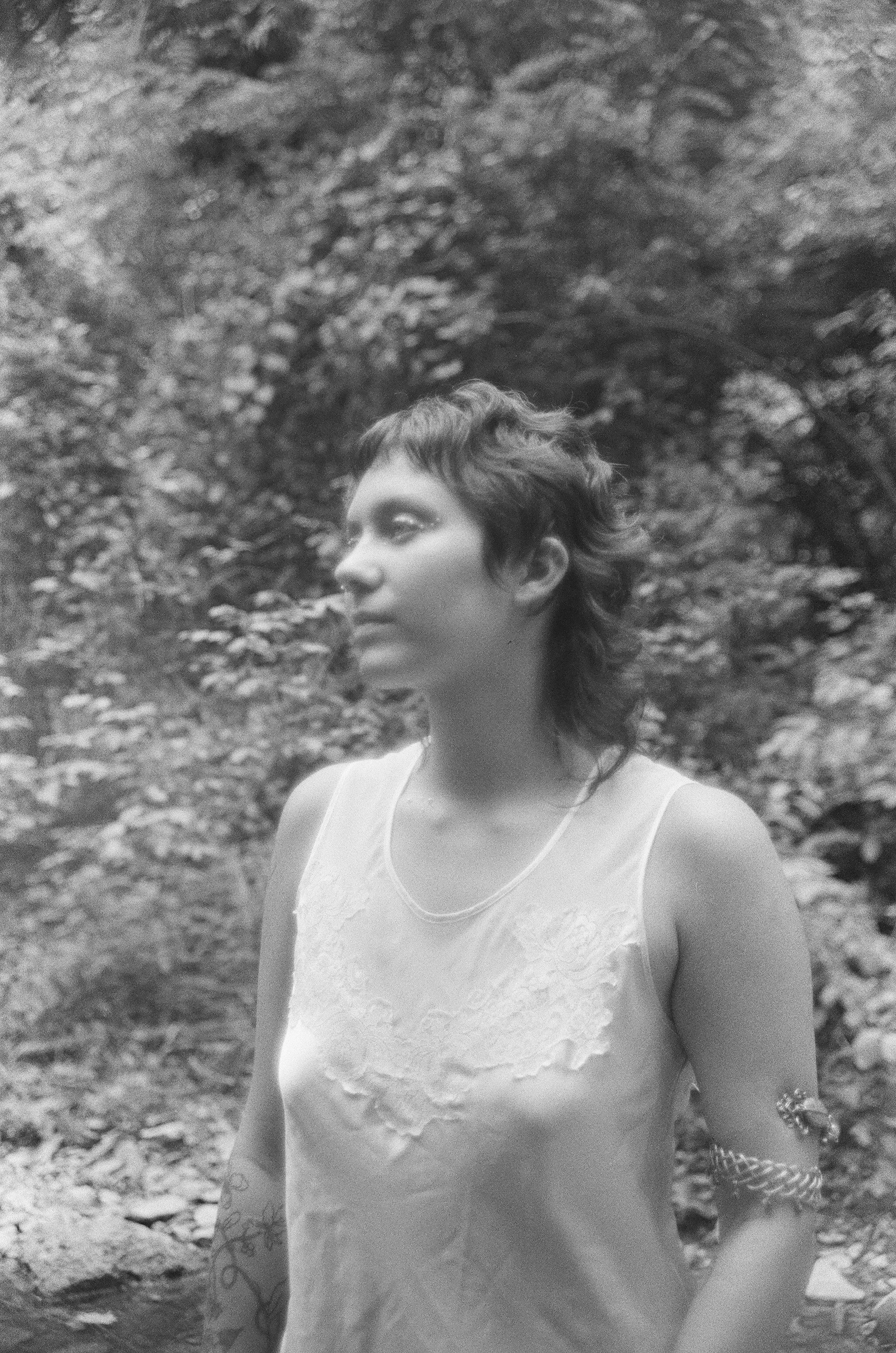 A woman with short, wavy hair standing outdoors near a wooded area, wearing a sleeveless top and jewelry, looking to the side.