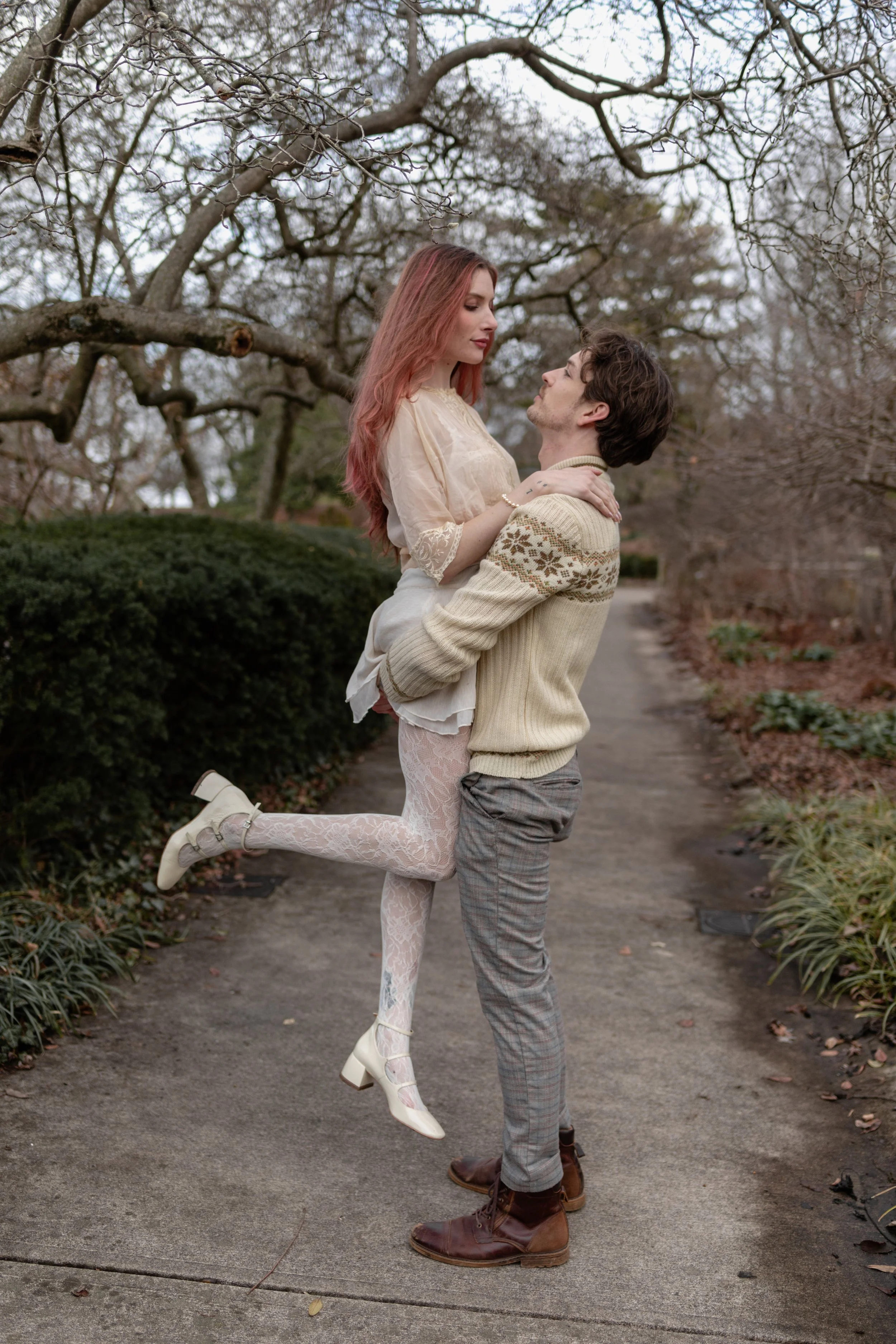 A woman with pink hair and a man with dark hair are outdoors during fall, with leafless trees and bushes in the background. The woman is wearing a cream-colored dress, white lace tights, and cream-colored shoes with heels. She is being lifted by the 