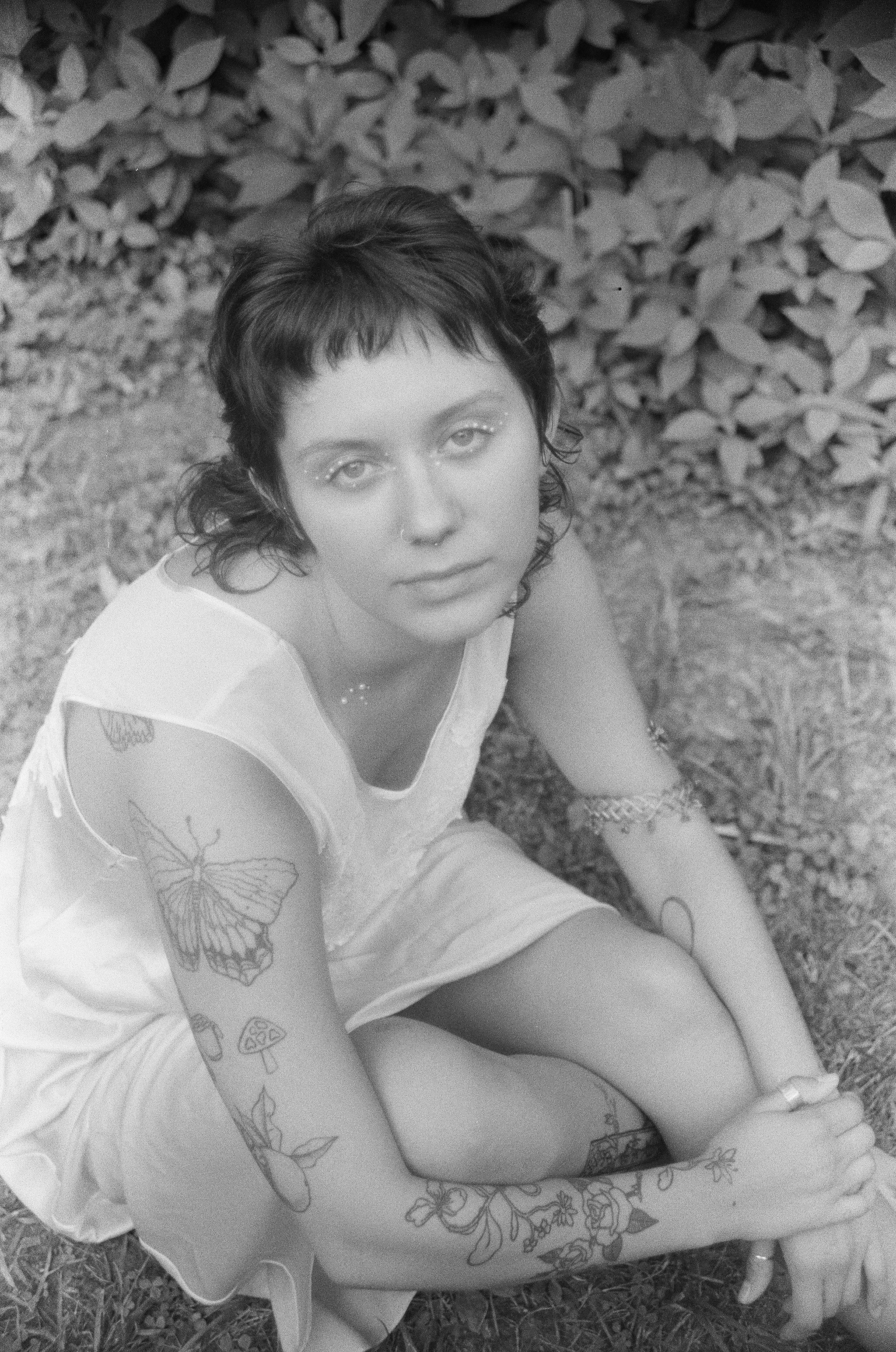 A woman with short, dark hair and tattoos on her arms, sitting outdoors on grass with a background of bushes, wearing a light-colored dress with flower and butterfly patterns, looking up at the camera.