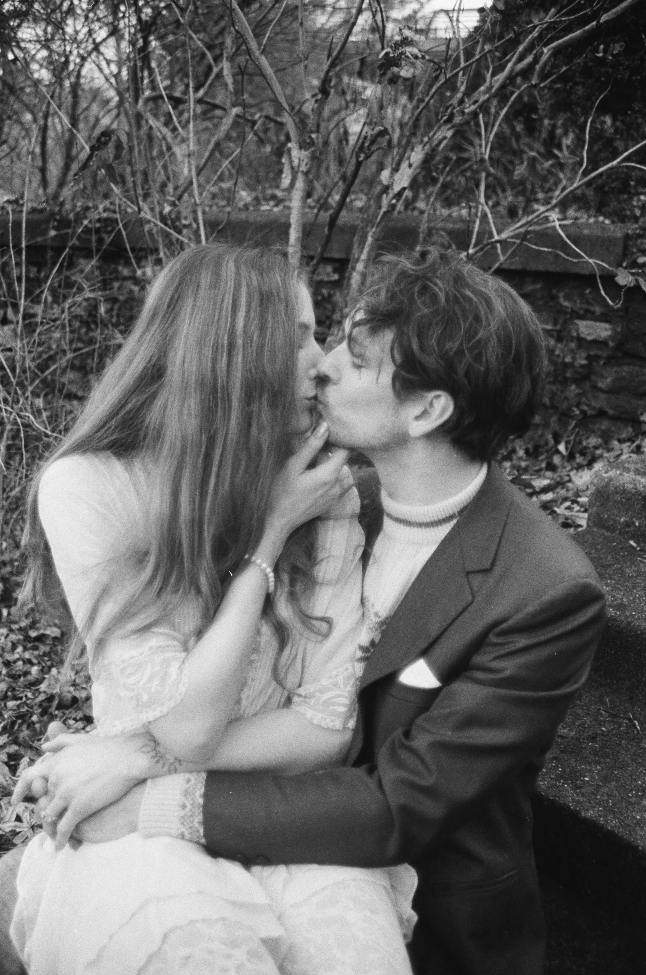 A black-and-white photo of a couple sharing a kiss outdoors, sitting close together with the woman's hand gently touching the man's face. The woman is wearing a light-colored dress with lace details, and the man is dressed in a dark suit with a shirt
