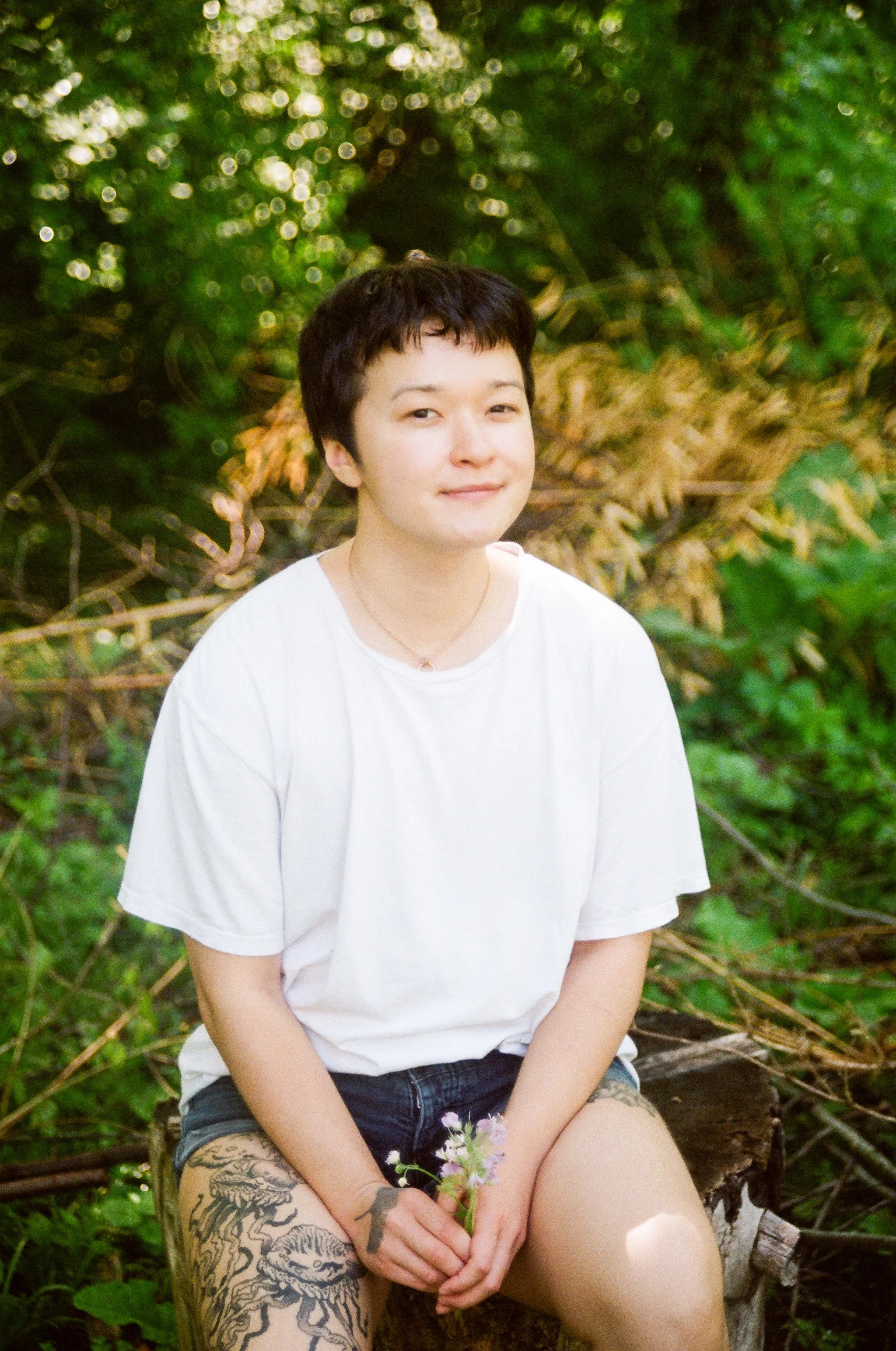 A young woman with short dark hair sitting outdoors on a log in a forested area, wearing a white t-shirt and dark shorts, holding a small bouquet of flowers, and smiling softly at the camera.