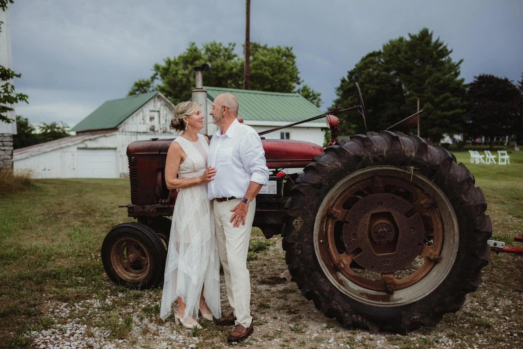 A couple dressed in white, standing close and smiling at each other, with old tractor and farmhouse in the background, on a grassy area with some trees and cloudy sky.