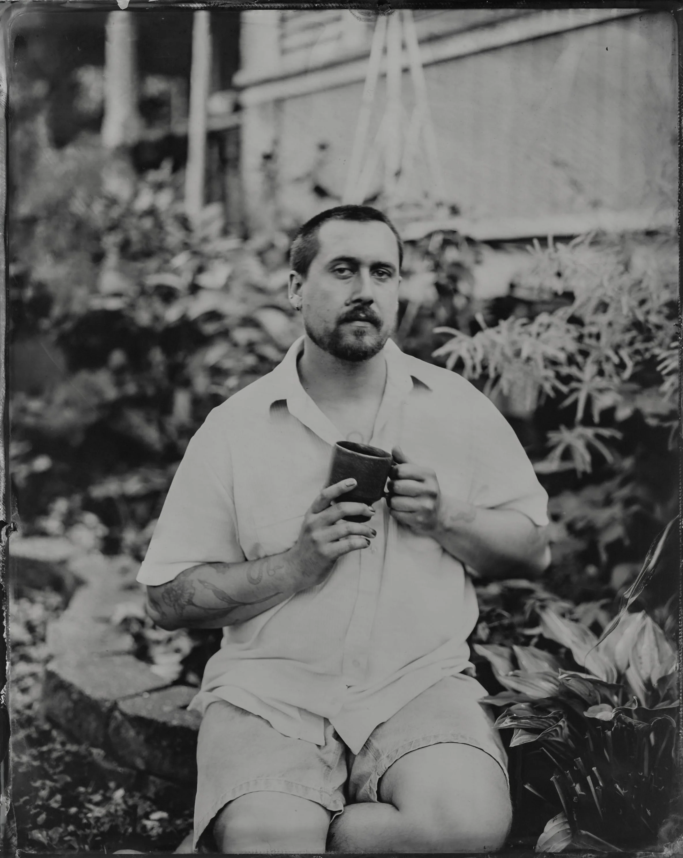 A black-and-white photo of a man sitting outdoors surrounded by plants and foliage, holding a cup with both hands, wearing a short-sleeved shirt and shorts, with tattoos visible on his arm and a contemplative expression.