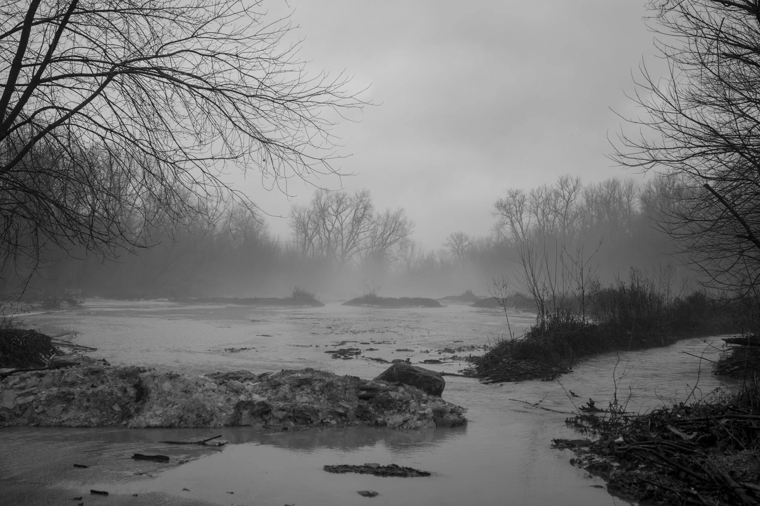 A black and white photo of a river with bare trees on both sides, some of which are reflected in the water. The scene appears foggy or overcast.