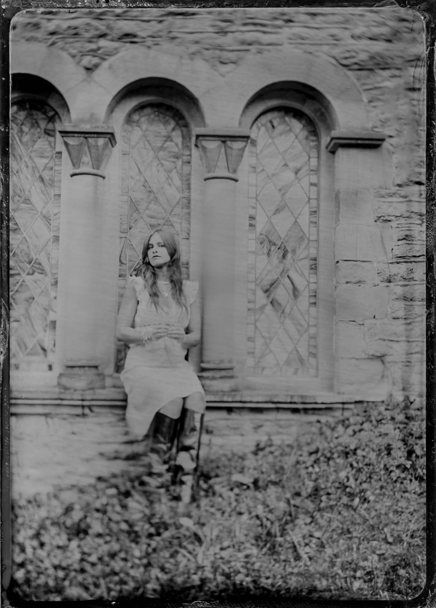Black and white photo of a young woman in a dress, standing in front of a brick wall with arched windows and decorative patterns, outdoors on a leaf-covered ground.