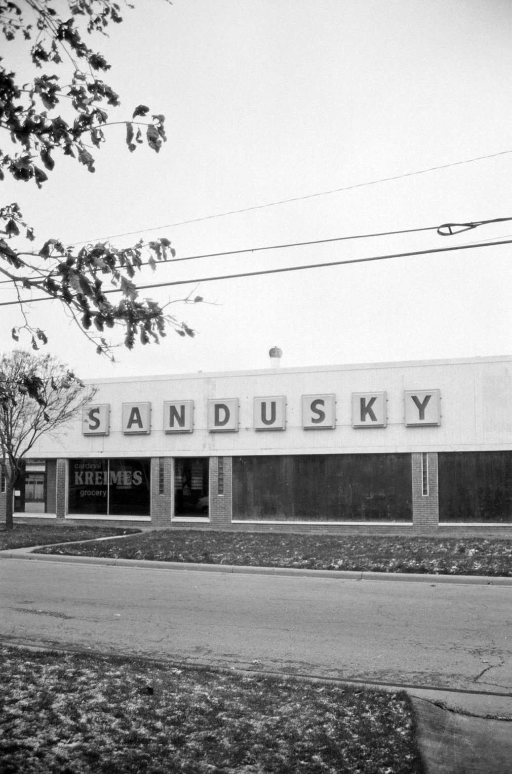 Black and white photo of a building with large mailbox tiles spelling SANDUSKY, a grocery store logo for KREMES, and a leafless tree in front.