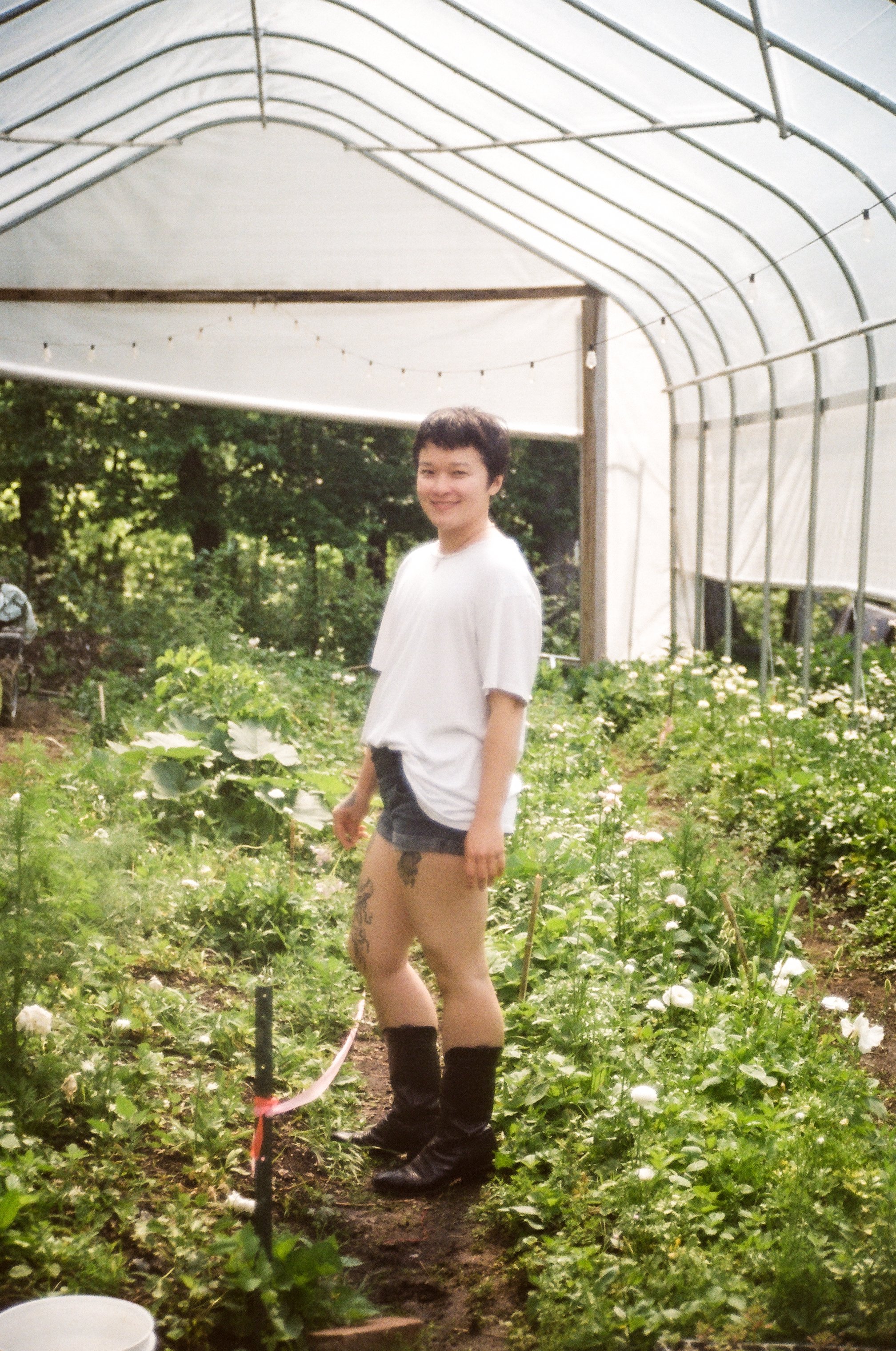 A person standing in a greenhouse surrounded by green plants and flowers, wearing black boots, shorts, and a white t-shirt, smiling at the camera.