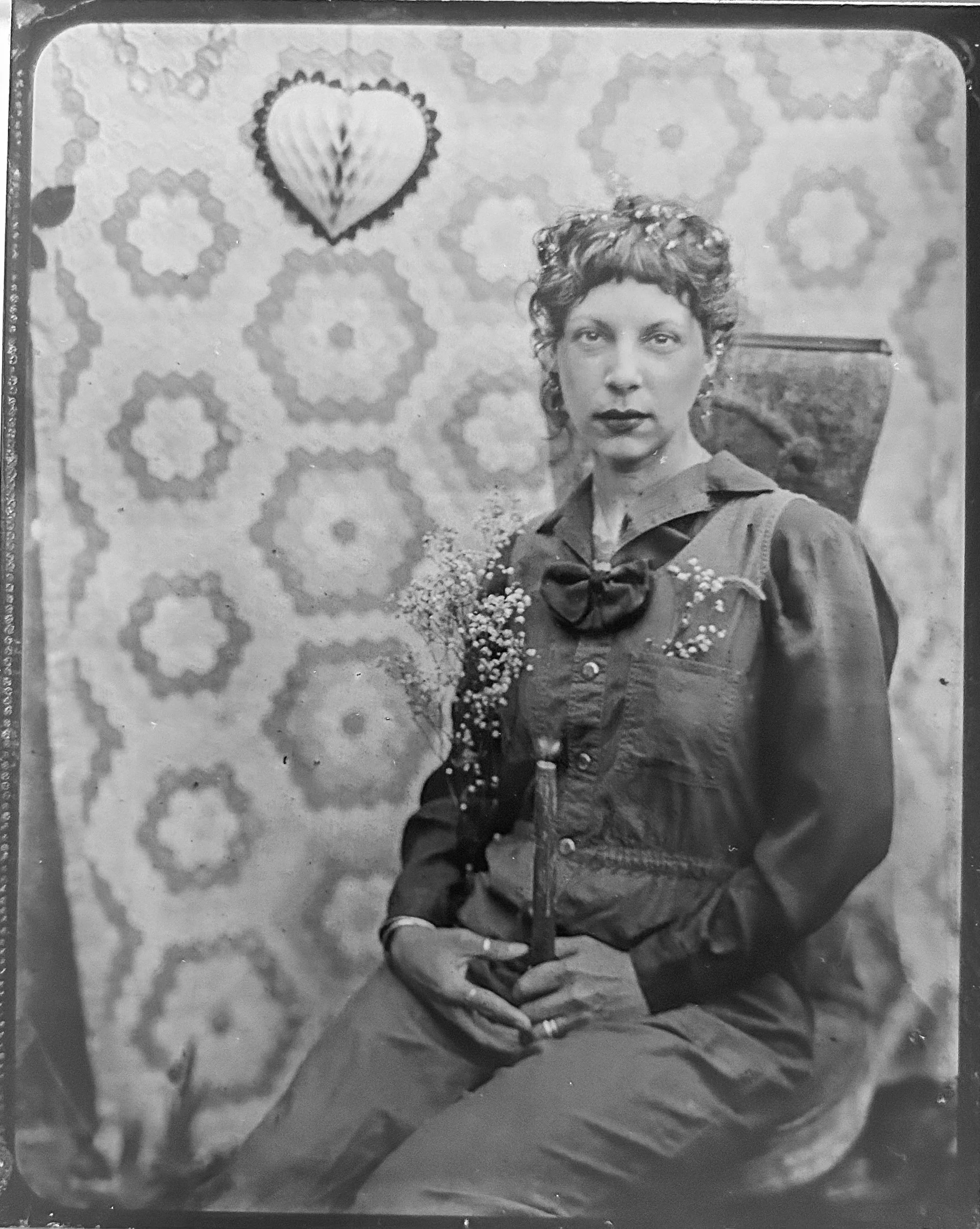 Black and white photograph of a woman with short curly hair, wearing a dark blouse with a bow at the collar, and sitting with her right hand resting on her hip. There is a large decorative heart-shaped object with a shell pattern above her right shou