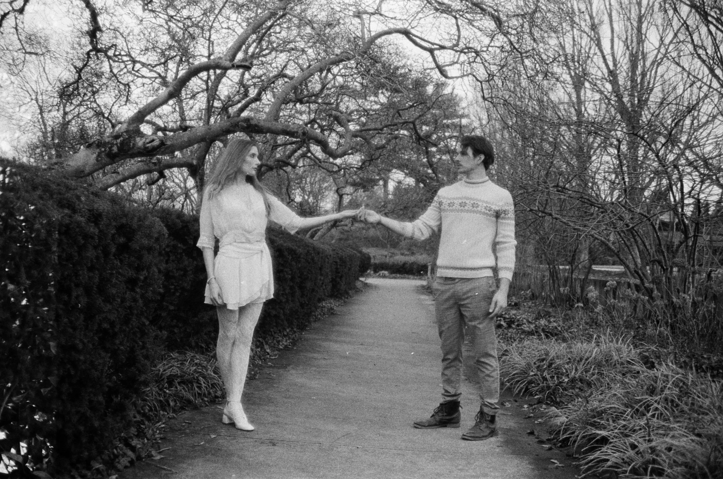 A black-and-white photo of a young woman and man holding hands while walking in a park with leafless trees and bushes, suggesting it is winter or late fall.