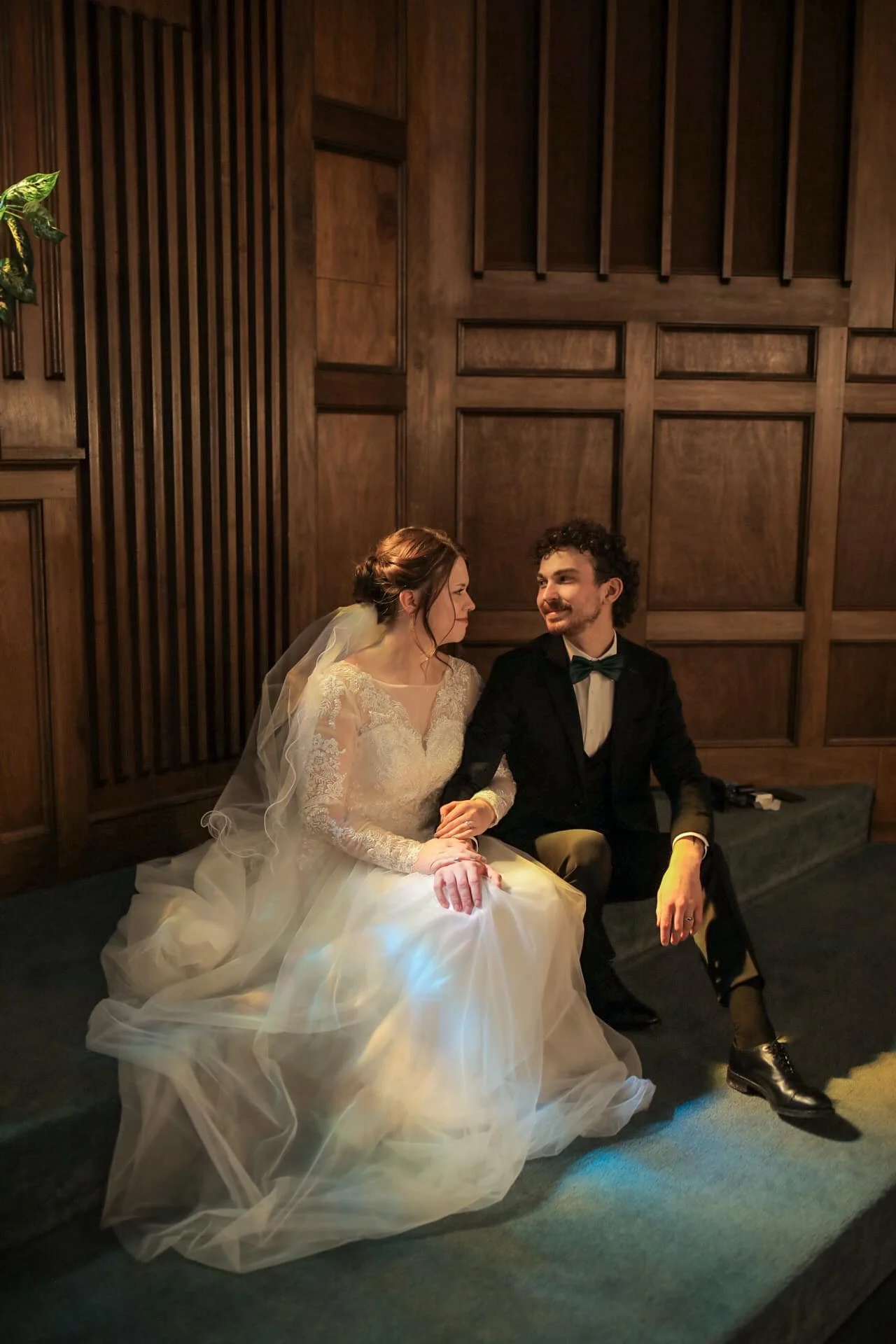 A bride and groom sitting close together in a wood-paneled room, smiling and looking at each other. The bride wears a wedding dress with lace details and a veil, while the groom is dressed in a tuxedo with a bow tie.