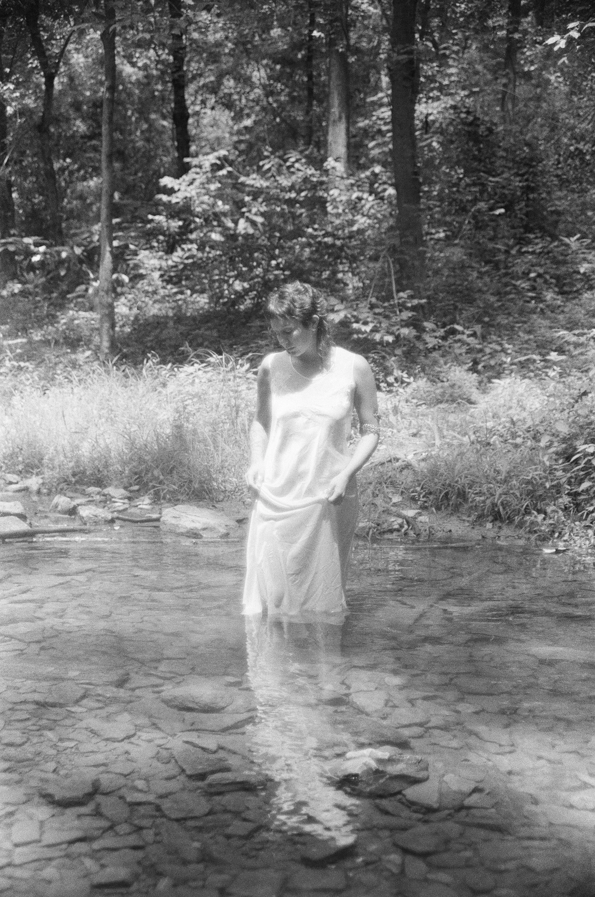 A young woman in a white dress standing in a shallow creek surrounded by large rocks and dense trees.