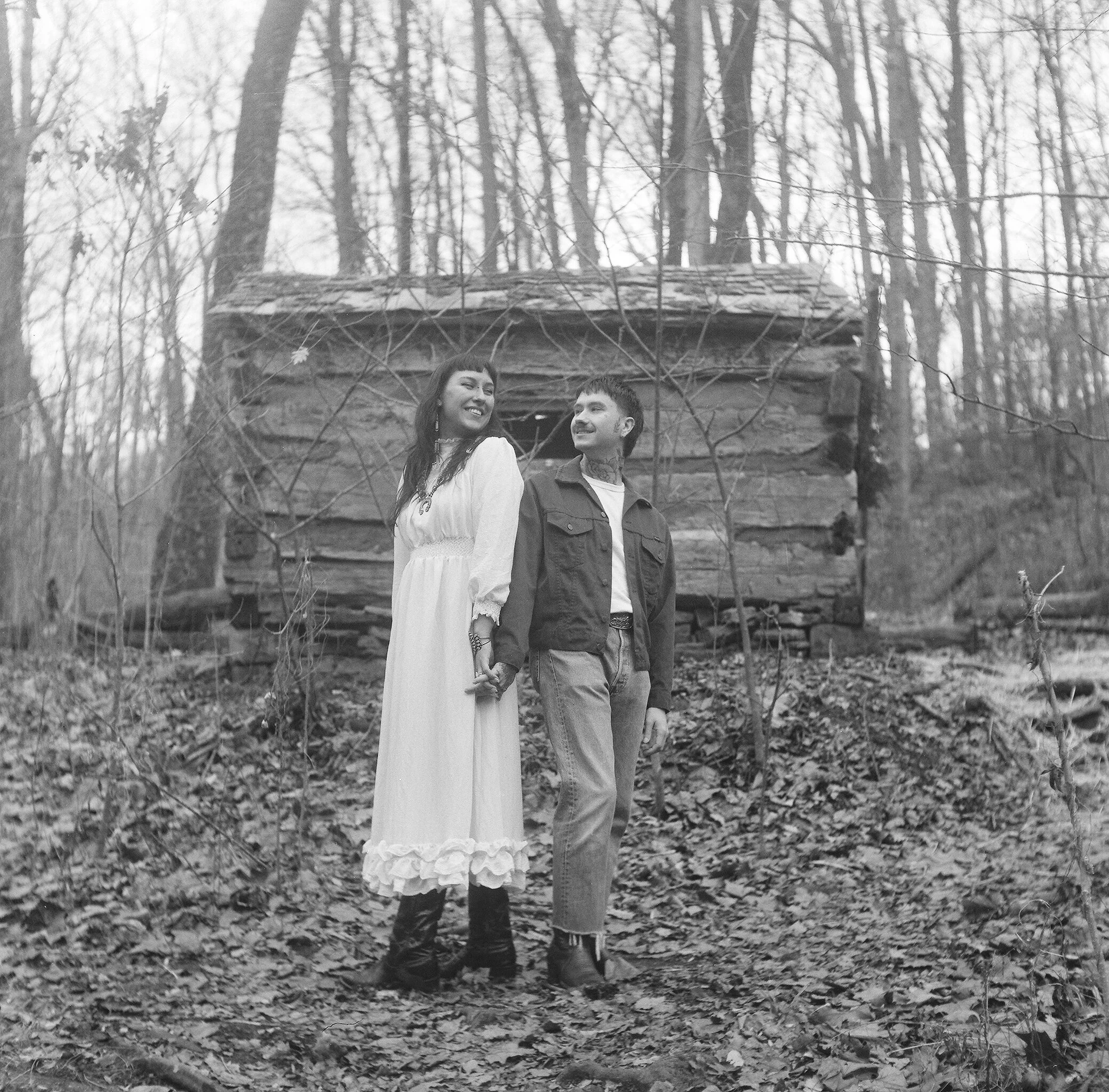 A smiling woman and man standing hand in hand outdoors in a forested area during daytime. Behind them is a wooden cabin among tall, leafless trees, with a ground covered in fallen leaves.
