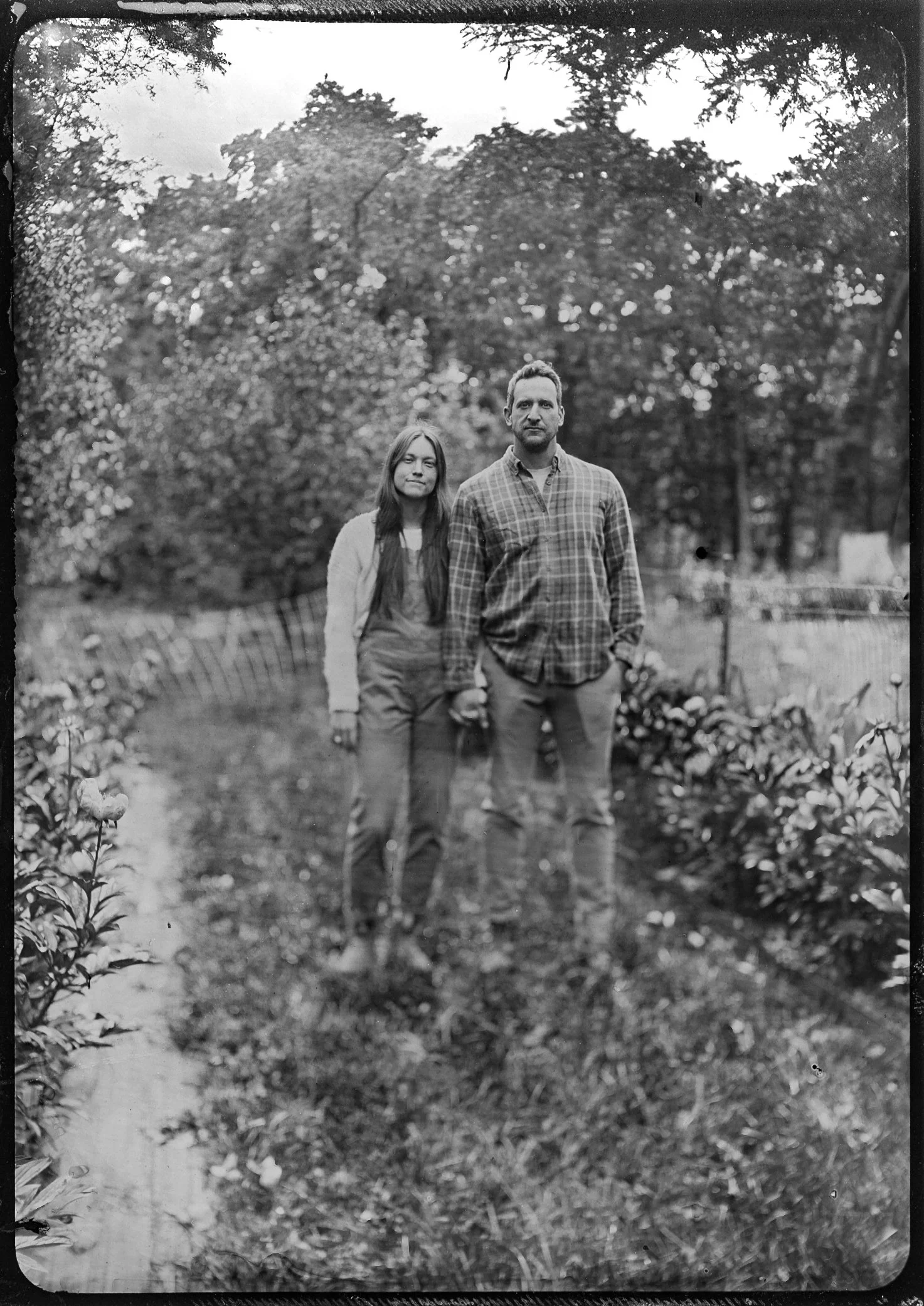 A black-and-white photo of a man and woman standing in a garden or outdoor area with trees and plants around them. They are posing for the camera, with the man in front and the woman slightly behind him.