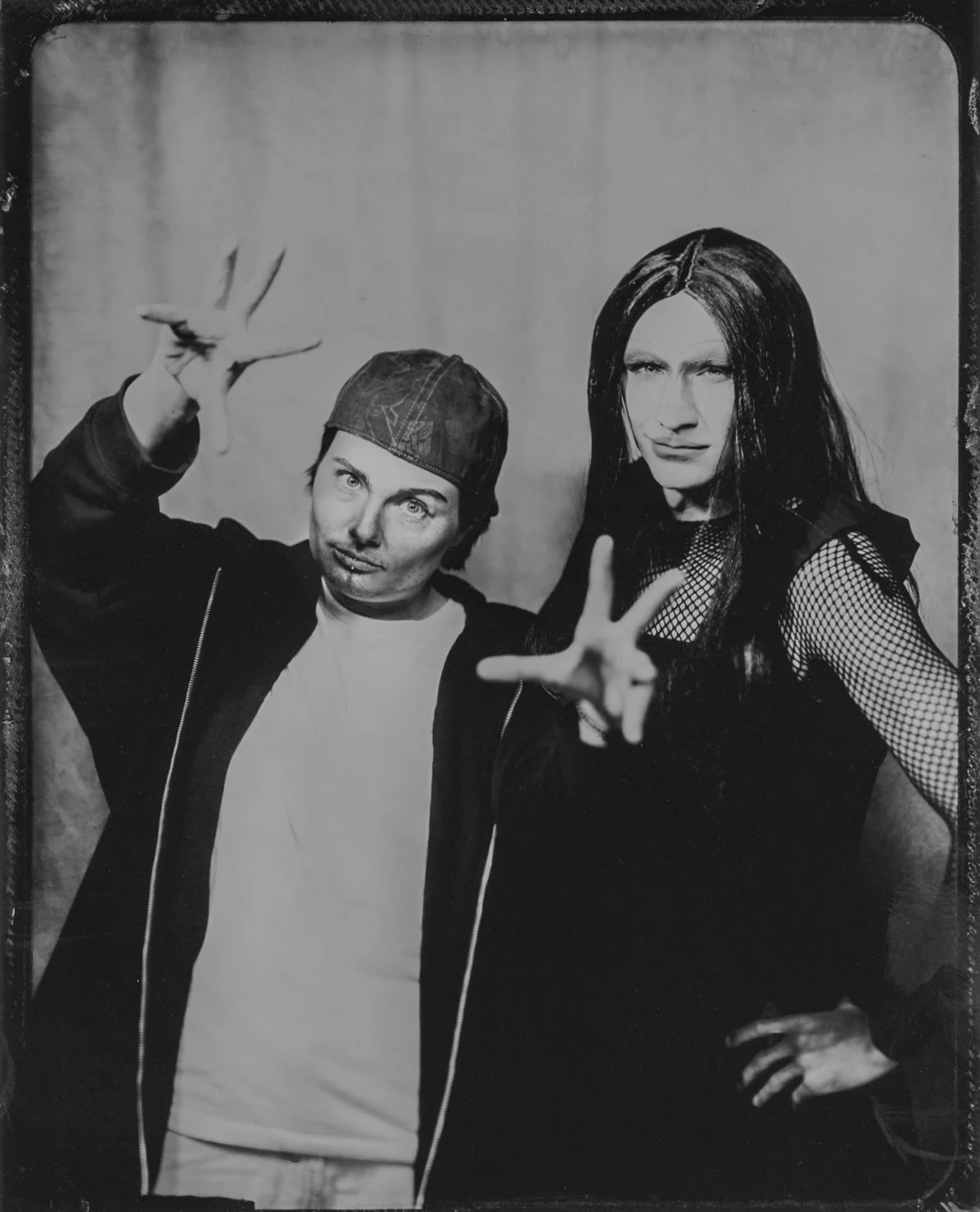 Black-and-white photo of two people making peace signs; person on left wears a cap and jacket, person on right has long dark hair and fishnet sleeves.