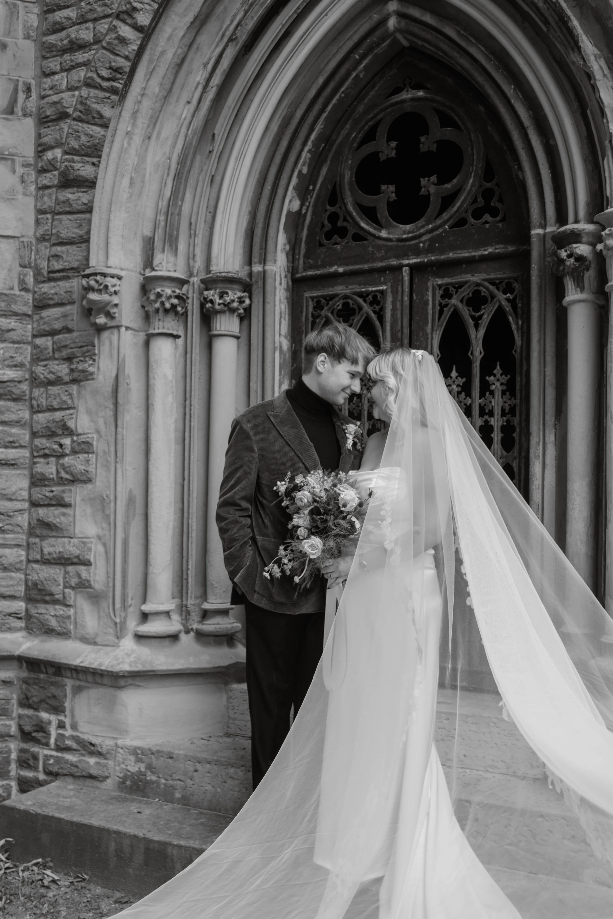 A black-and-white photo of a bride and groom standing close to each other outside a stone building with gothic windows, with their foreheads touching.