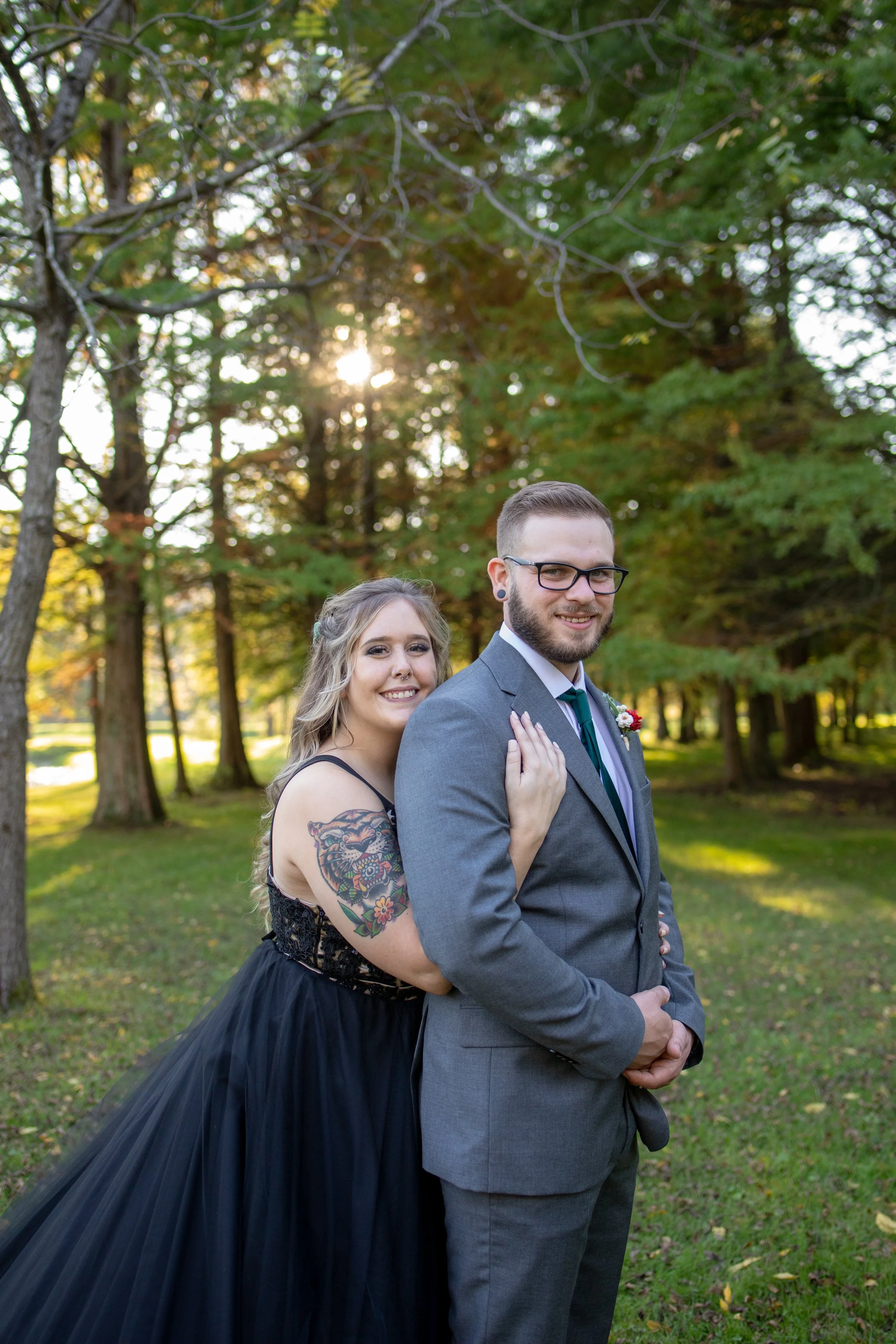 A smiling couple embracing outdoors in a park with trees in the background, the woman has long hair with tattoos on her arm, wearing a black dress, and the man is in a gray suit with glasses and a boutonniere.