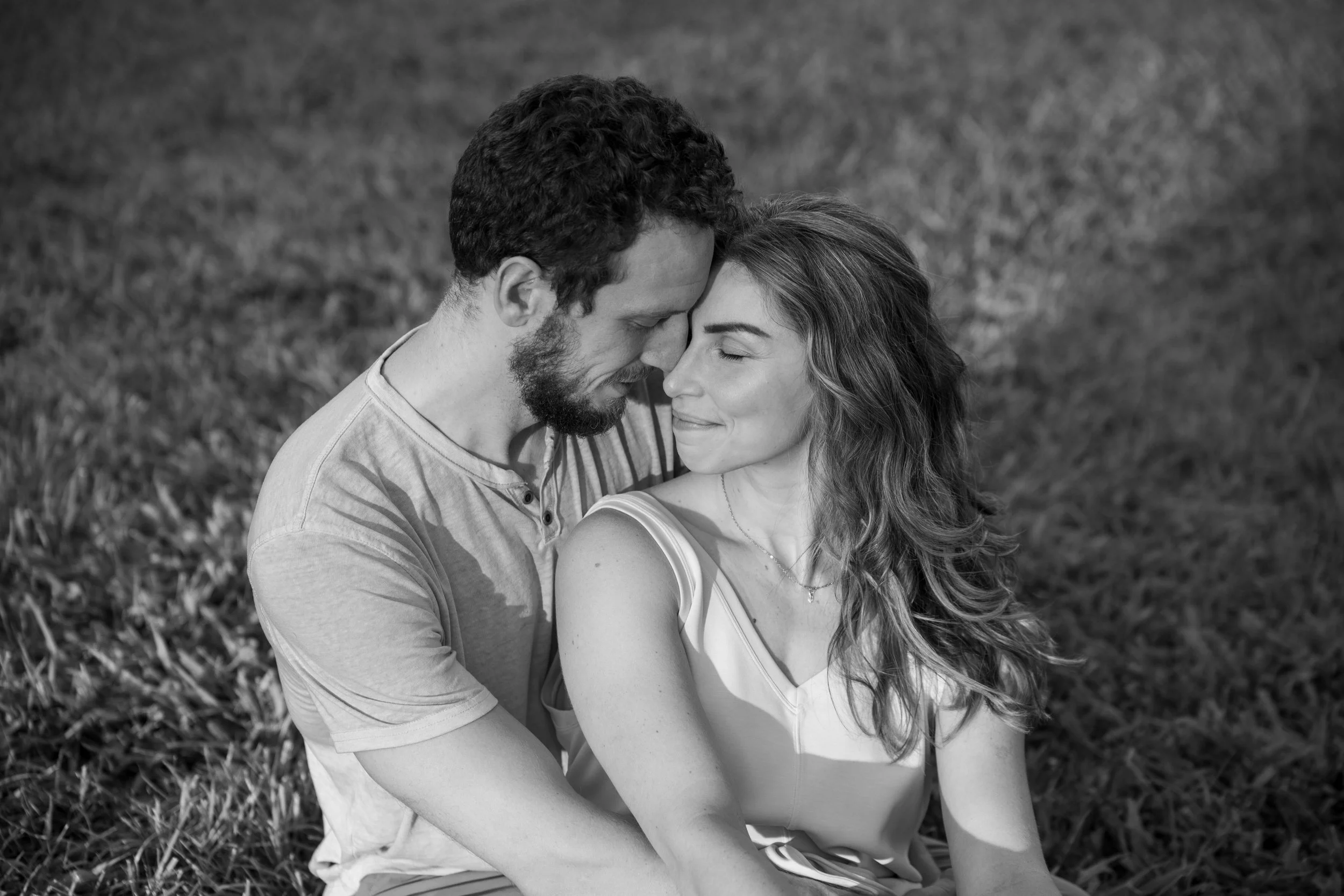 A black and white photograph of a couple sitting on the grass, touching foreheads with eyes closed, smiling softly, in a peaceful outdoor setting.