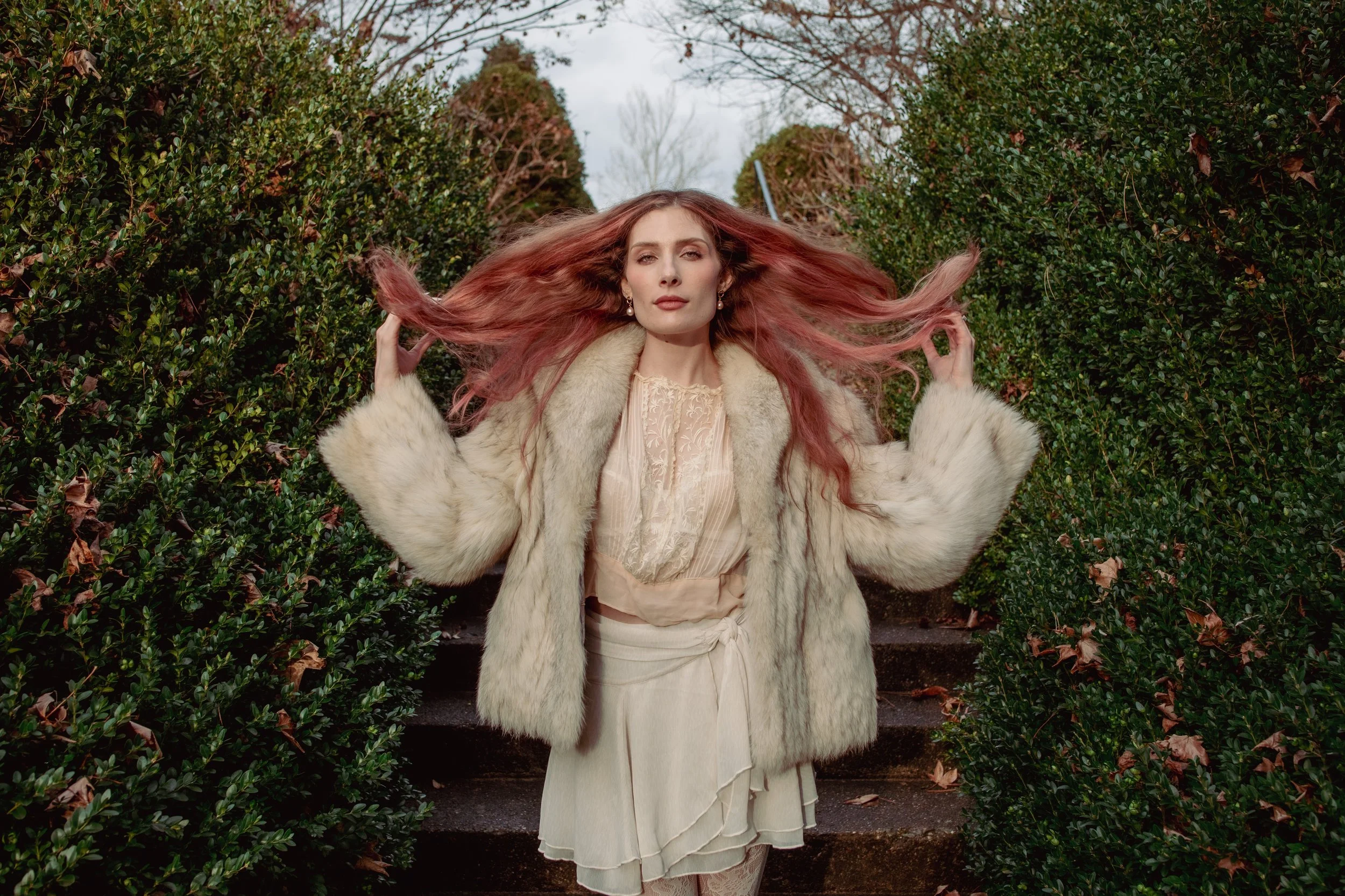 A woman standing on outdoor steps between bushes, with her hair spread out as she holds it, wearing a fur coat over a dress.