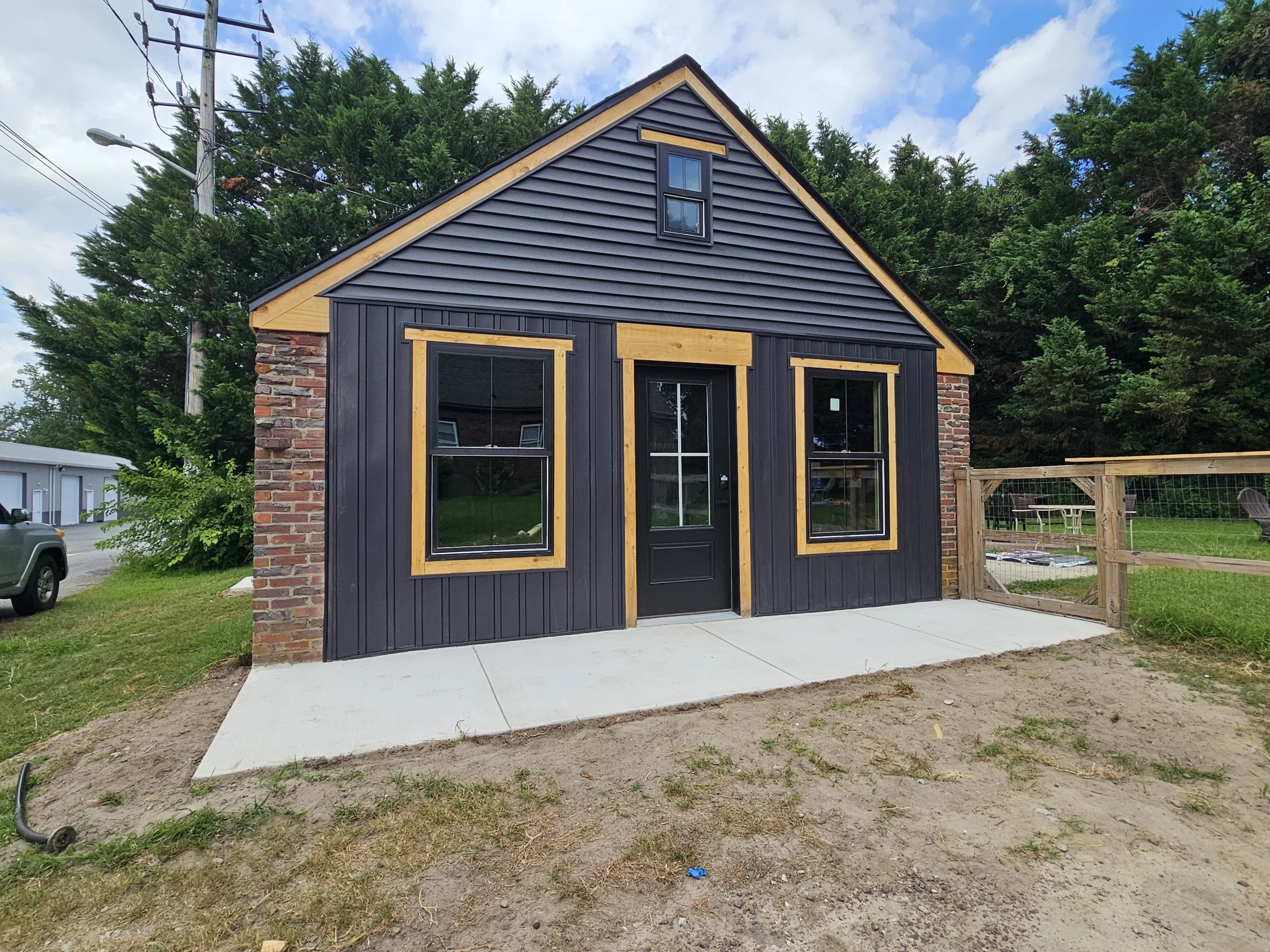 Exterior of a converted garage with new dark siding, original brick accents, and new natural wood trim, under a partly cloudy sky, with a concrete sidewalk in front and a fenced backyard to the side.