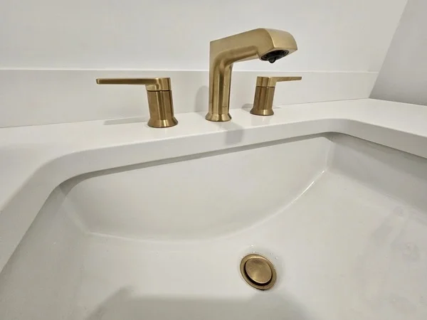 Close-up of a white bathroom sink with a gold faucet and handles.
