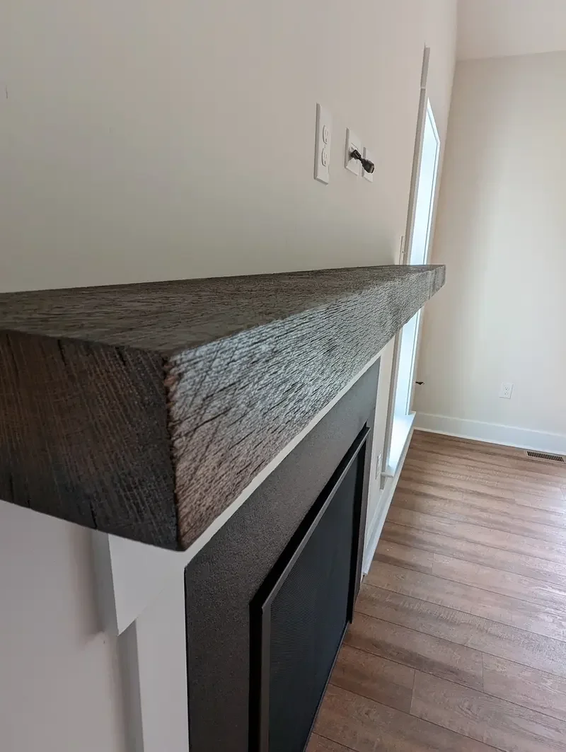 Close-up of a wooden fireplace mantel with a textured dark finish in a room with hardwood floors and electrical outlets.