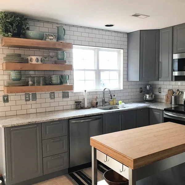 Full kitchen renovation. A modern kitchen with gray cabinets, white subway tile backsplash, and a window above the sink. Open wooden shelves hold dishes and decorative items. A wooden island is in the foreground. Milton, Delaware