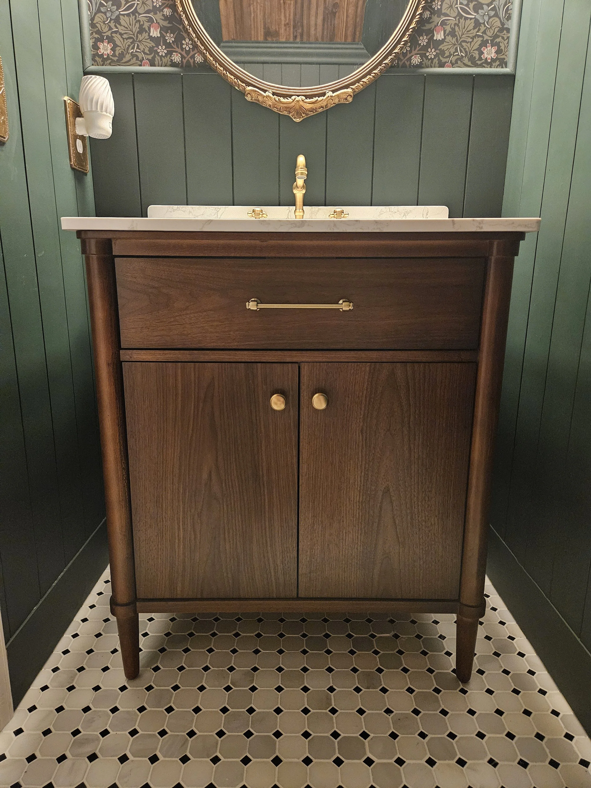 A vintage wooden bathroom vanity with a marble countertop, a brass faucet, two small cabinet doors with brass handles, and a large round mirror with an ornate gold frame. The vanity is placed against a dark paneled wall with floral wallpaper above, and it stands on a black and white hexagonal tile floor.