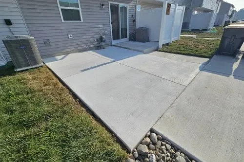New concrete patio in backyard with a grassy area, sliding glass door, and a small section of rocks.