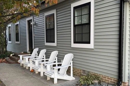 Four white Adirondack chairs lined up outside a gray house with four black-framed windows, green plants, and a sidewalk.